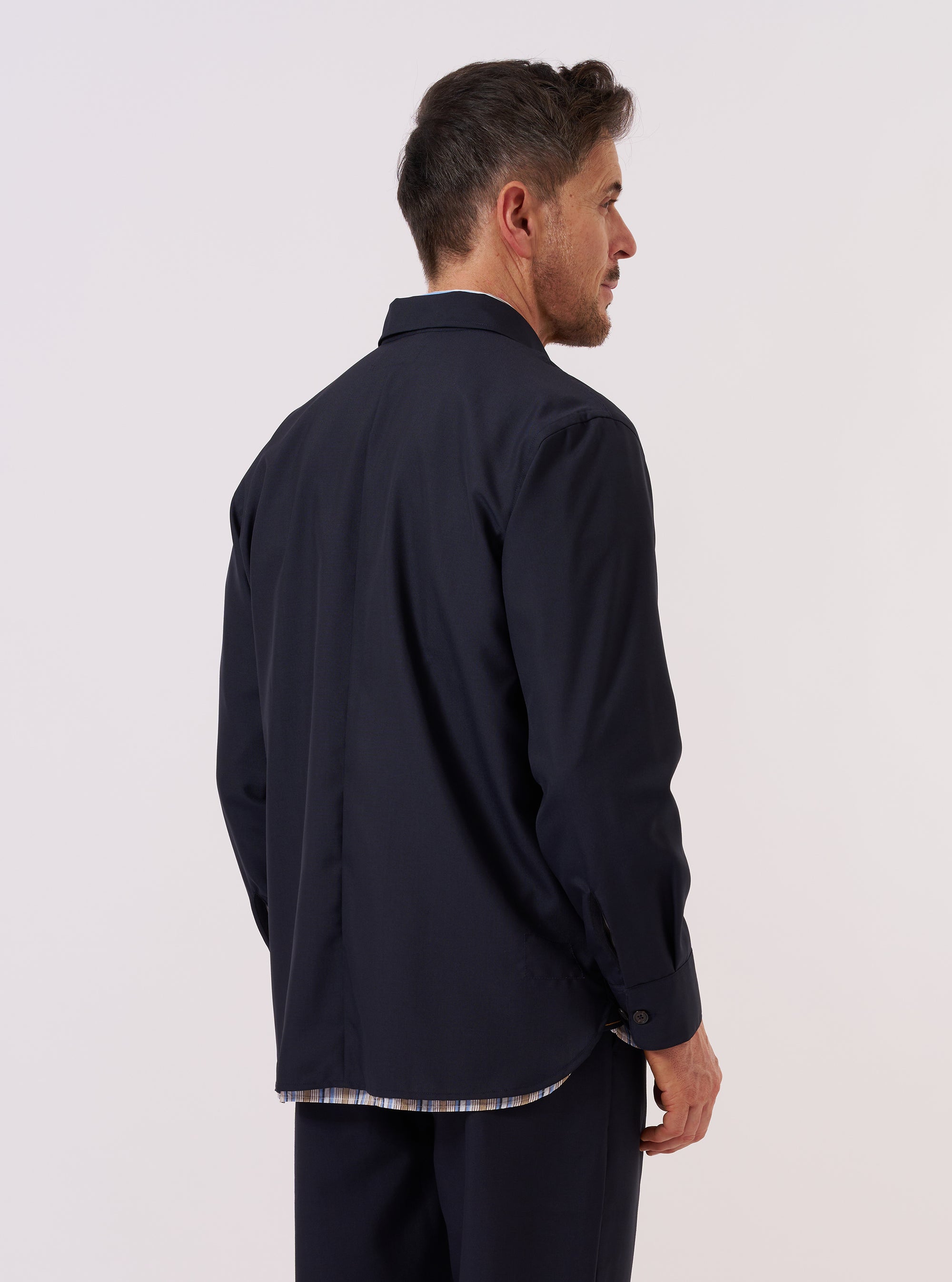 A model is wearing the Navy Tropical Suiting Travail Overshirt, shown from a side-angle view against a plain white background. The overshirt features a classic collar, a button-down front, and a large patch pocket visible on the side. The garment is constructed from a smooth, navy blue tropical suiting fabric.