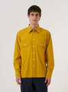 A model wears the Golden Brown Organic Fine Poplin CPO Shirt over a neutral-colored top, standing against a plain, light-colored background. The shirt features a pointed collar, a full button-down front, and two large buttoned patch pockets on the chest. The organic cotton poplin fabric has a smooth, crisp appearance in a rich golden brown hue.
