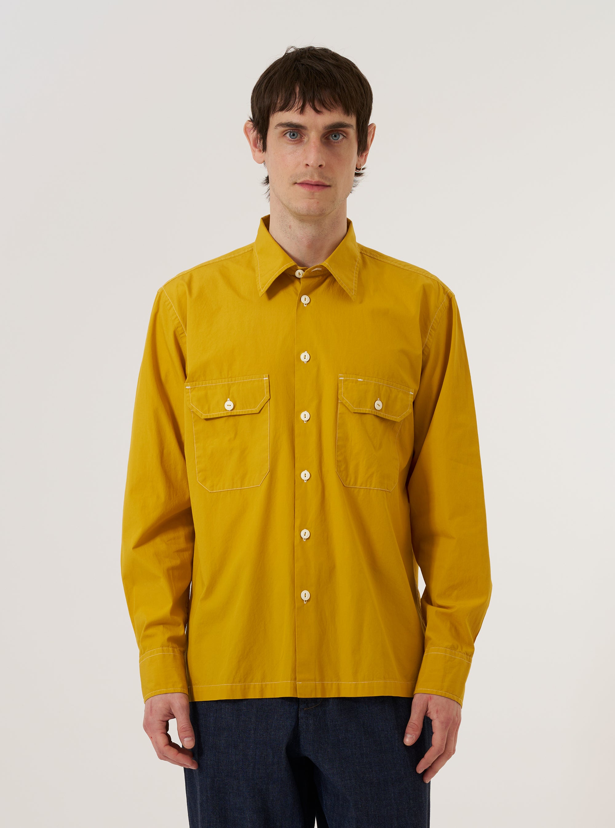 A model wears the Golden Brown Organic Fine Poplin CPO Shirt over a neutral-colored top, standing against a plain, light-colored background. The shirt features a pointed collar, a full button-down front, and two large buttoned patch pockets on the chest. The organic cotton poplin fabric has a smooth, crisp appearance in a rich golden brown hue.