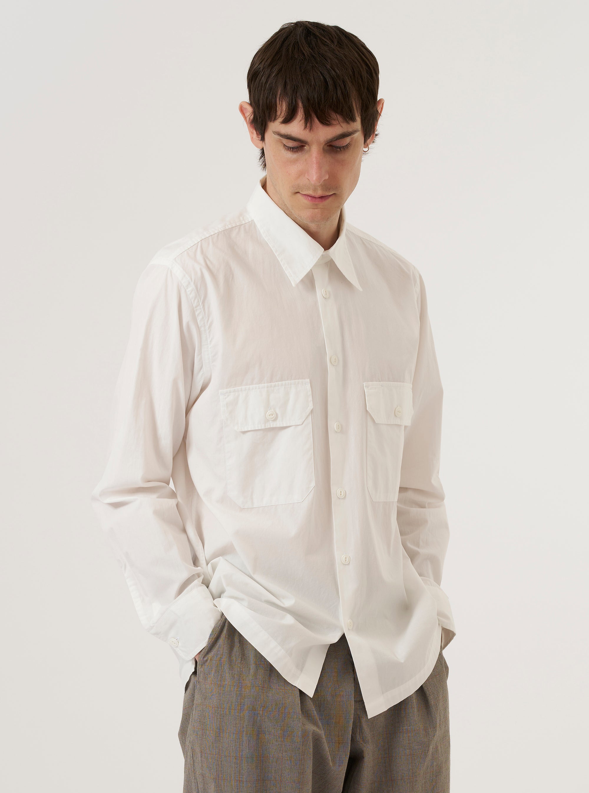 A model wears the Ecru Organic Fine Poplin CPO Shirt over a dark-colored top, standing against a plain, neutral studio background. The shirt features a pointed collar, a central button-down closure, and two large buttoned patch pockets on the chest. The ecru-colored organic cotton poplin fabric has a smooth, fine texture and a clean, utility-inspired appearance.