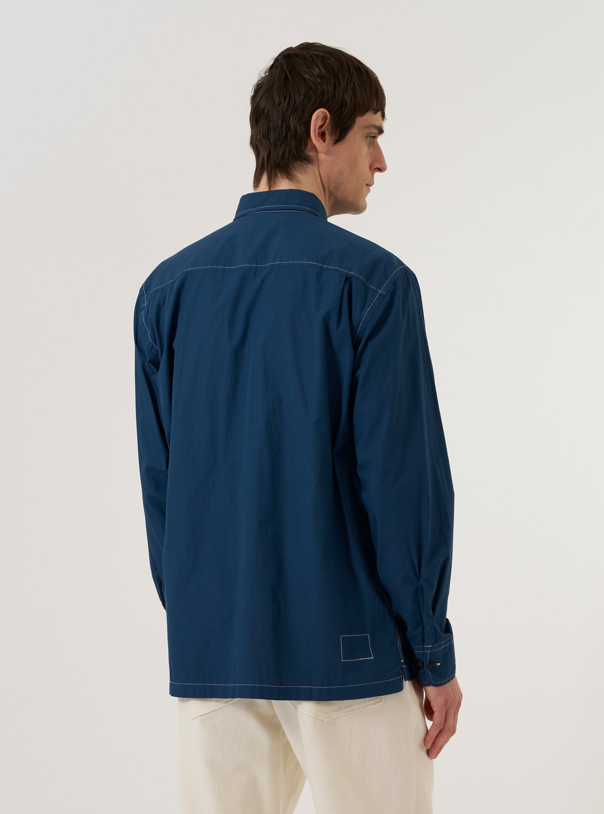 A model wears the Navy Organic Fine Poplin CPO Shirt, shown from a rear three-quarter angle against a plain white background. The image highlights the shirt's relaxed fit, simple shoulder yoke, and the smooth, crisp texture of the dark navy organic cotton poplin fabric. The garment features a straight hem and a structured collar.
