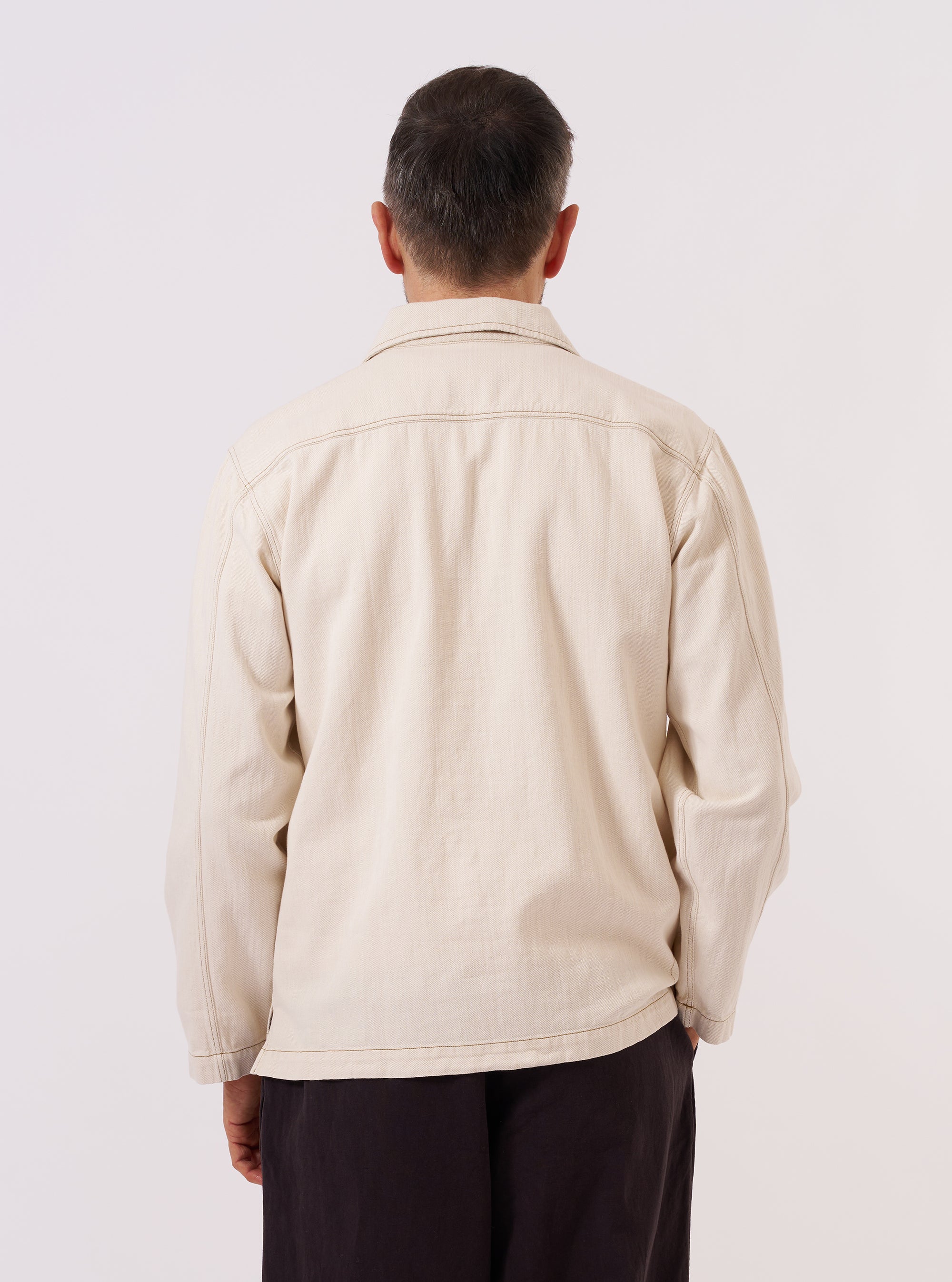 A model wears the Ecru Koko Cotlin Smock Overshirt, shown from a rear three-quarter angle against a plain, neutral background. The pullover-style garment features a relaxed fit, a straight hem, and a simple collar. The ecru fabric exhibits a natural, textured appearance characteristic of a cotton and linen blend.