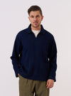 A model wears the Indigo Recycled Herringbone Smock Overshirt against a plain, neutral studio background. The overshirt features a pullover smock design with a buttoned half-placket, a structured collar, and three large patch pockets on the front. The dark indigo fabric displays a distinct herringbone weave texture throughout.
