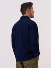 A model is wearing the Indigo Recycled Herringbone Smock Overshirt, shown from a side angle against a plain, neutral studio background. The overshirt features a pullover smock design with a relaxed fit and a deep indigo blue color. The fabric displays a distinct herringbone weave texture, and the garment is finished with a simple pointed collar and a straight hem.