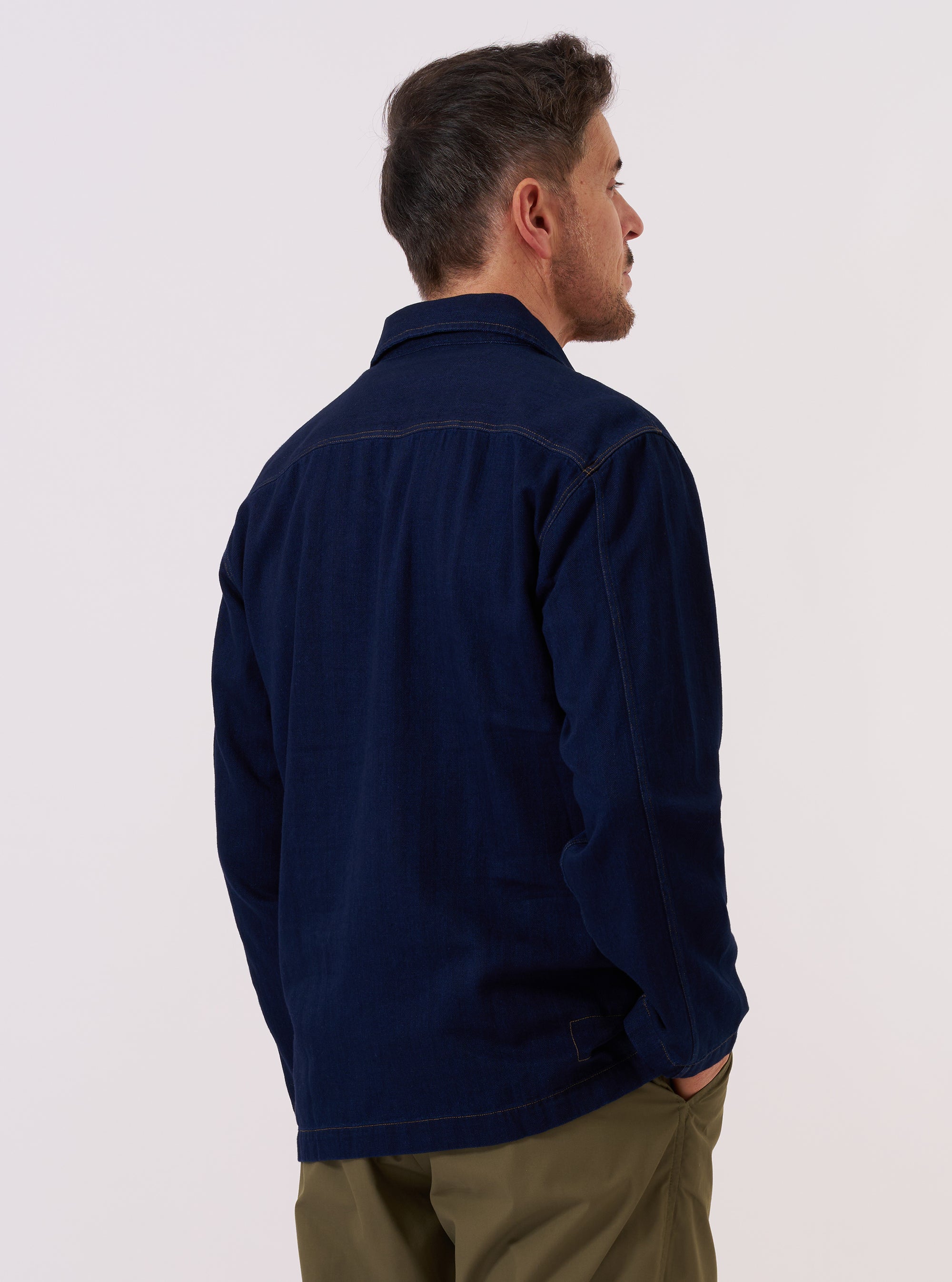 A model is wearing the Indigo Recycled Herringbone Smock Overshirt, shown from a side angle against a plain, neutral studio background. The overshirt features a pullover smock design with a relaxed fit and a deep indigo blue color. The fabric displays a distinct herringbone weave texture, and the garment is finished with a simple pointed collar and a straight hem.