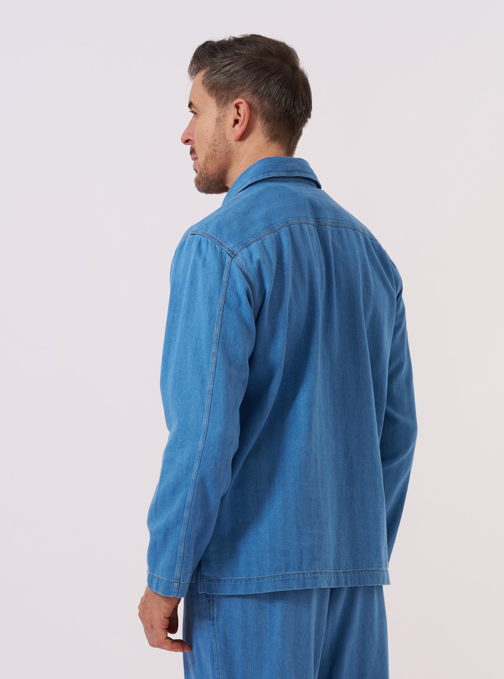 A model wears the Washed Indigo Recycled Herringbone Smock Overshirt, shown from the back against a plain, neutral background. The overshirt features a relaxed pullover fit, a simple collar, and a straight hem. The washed indigo fabric has a visible herringbone texture.