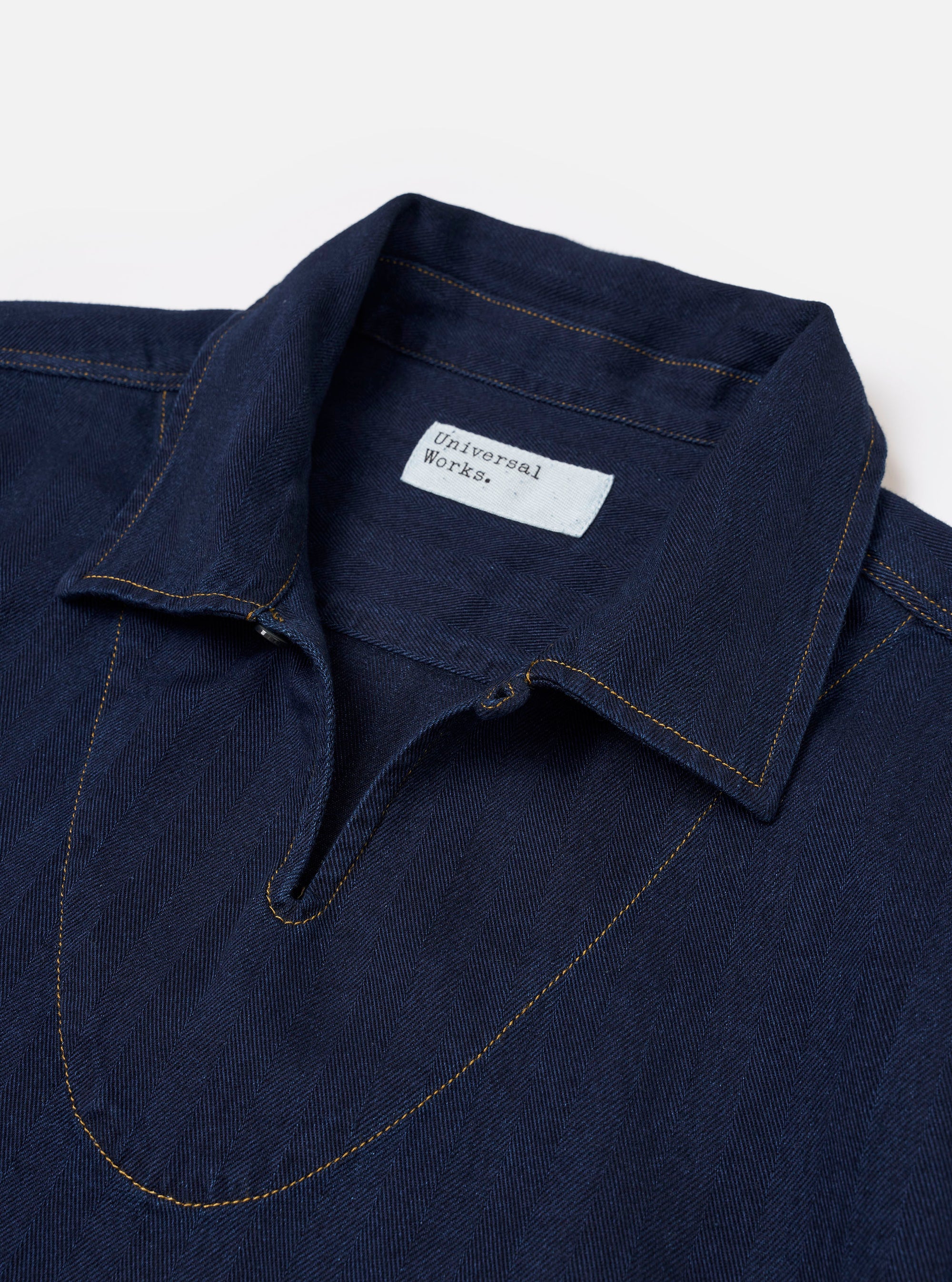 The Indigo Recycled Herringbone Smock Overshirt is shown from the back as a flat lay against a plain white background. The image highlights the garment's textured herringbone weave in a deep indigo blue, featuring a simple, straight-cut back panel and a clean hemline.