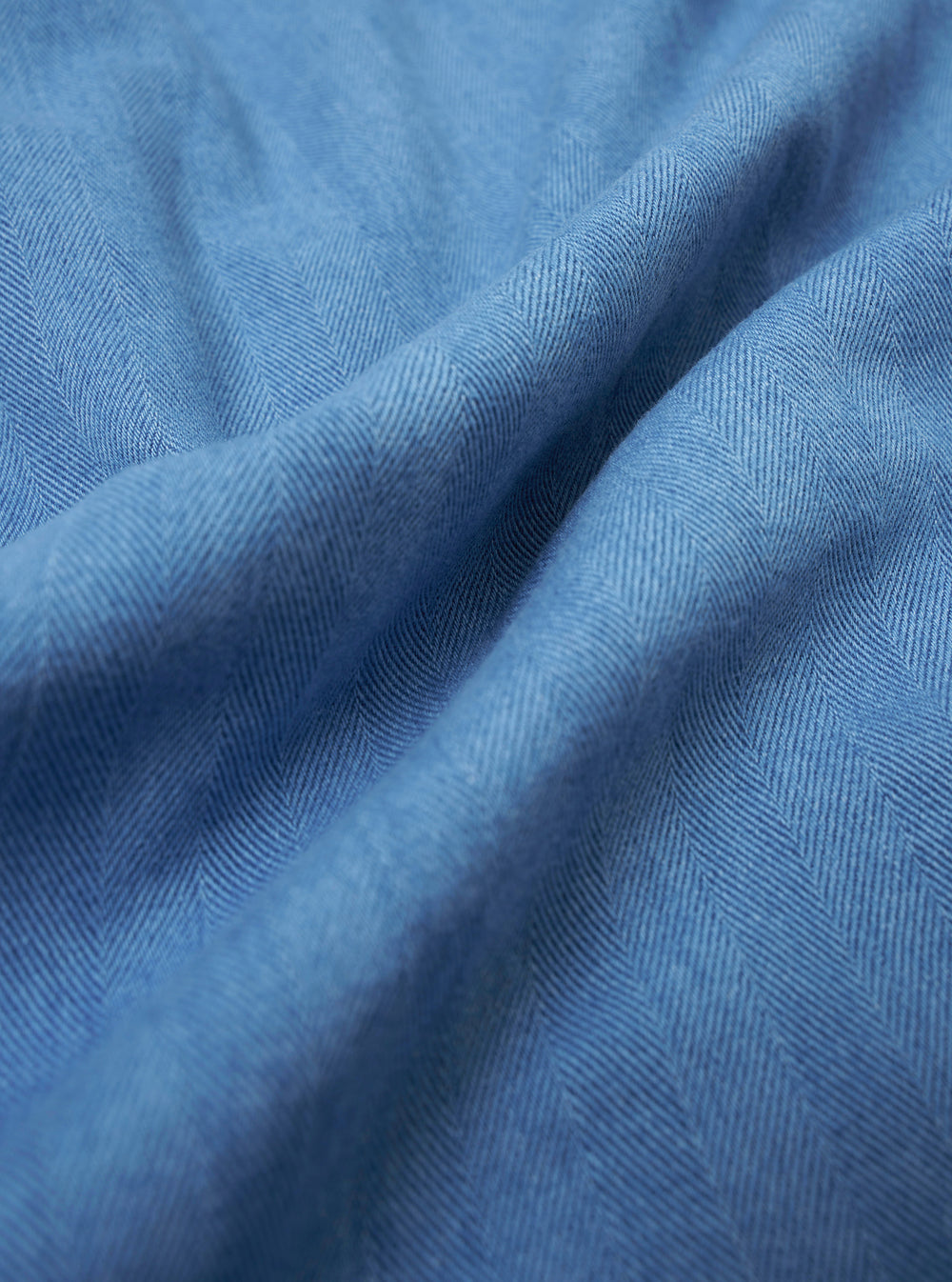 A close-up fabric swatch of the Washed Indigo Recycled Herringbone Smock Overshirt highlights the intricate V-pattern of the herringbone weave. The image displays the rich, varied blue tones of the indigo dye and the textured appearance of the recycled cotton and wool blend.