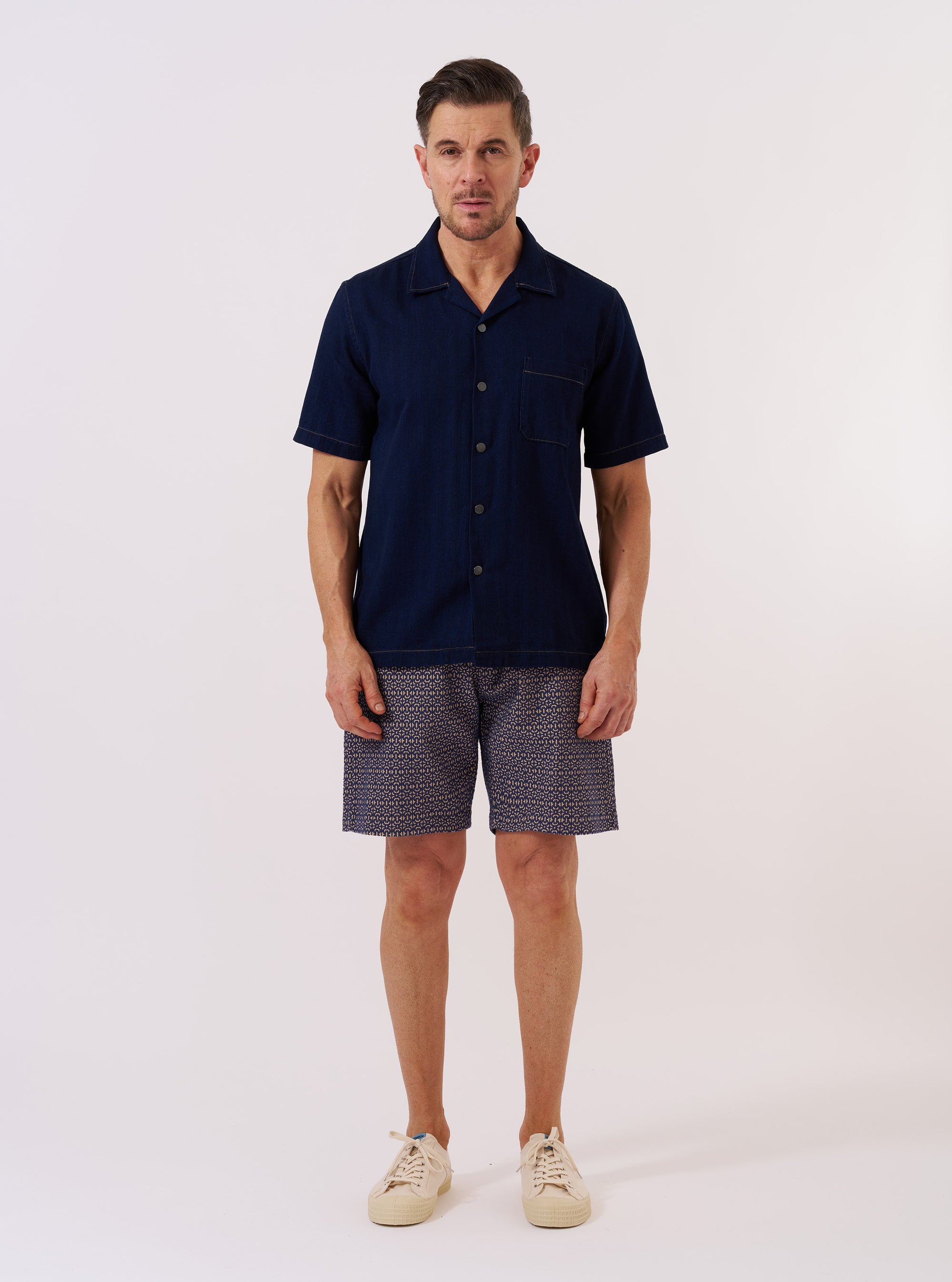 The Indigo Recycled Herringbone Road Shirt is shown as a flat lay against a white background. The shirt features a camp collar, a single chest patch pocket, and a straight hem. The indigo blue fabric has a visible herringbone weave pattern throughout.