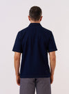 A model wears the Indigo Recycled Herringbone Road Shirt, shown from the back against a plain white studio background. The shirt features a relaxed fit, short sleeves, and a straight hem. The indigo blue fabric has a visible herringbone weave texture. No mannequin is present in the image.