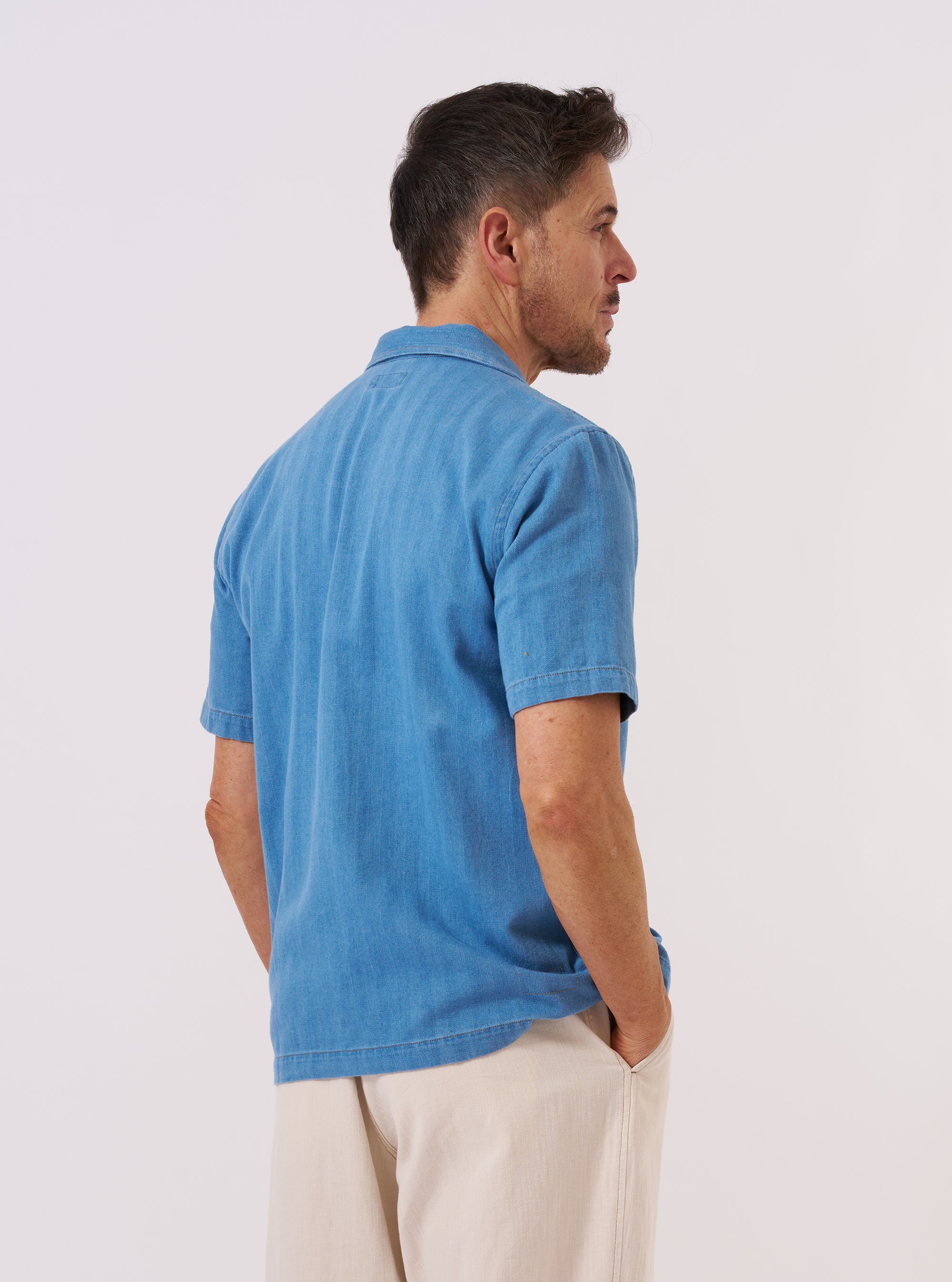 A model is wearing the Washed Indigo Recycled Herringbone Road Shirt, shown from the back against a plain white background. The shirt features a relaxed fit, a straight hem, and a simple back yoke. The washed indigo blue fabric has a visible herringbone weave texture throughout. No mannequin is present in the image.