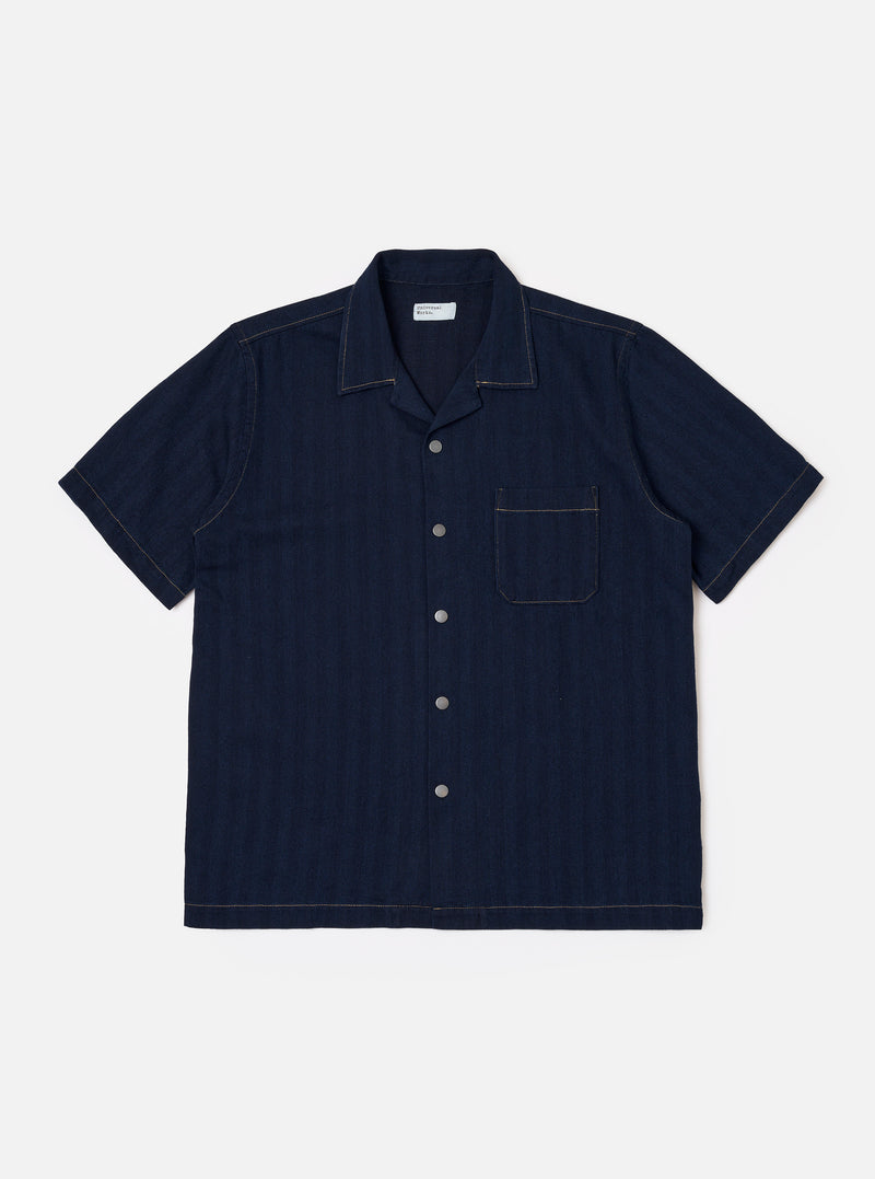 The Indigo Recycled Herringbone Road Shirt is shown as a flat lay against a white background. The short-sleeve shirt features a camp collar, a full button-front placket with brown corozo buttons, and three front patch pockets: one at the left chest and two at the waist. The fabric is made from indigo-dyed recycled cotton with a distinct herringbone weave.