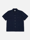 The Indigo Recycled Herringbone Road Shirt is shown as a flat lay against a white background. The short-sleeve shirt features a camp collar, a full button-front placket with brown corozo buttons, and three front patch pockets: one at the left chest and two at the waist. The fabric is made from indigo-dyed recycled cotton with a distinct herringbone weave.