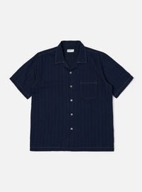 The Indigo Recycled Herringbone Road Shirt is shown as a flat lay against a white background. The short-sleeve shirt features a camp collar, a full button-front placket with brown corozo buttons, and three front patch pockets: one at the left chest and two at the waist. The fabric is made from indigo-dyed recycled cotton with a distinct herringbone weave.