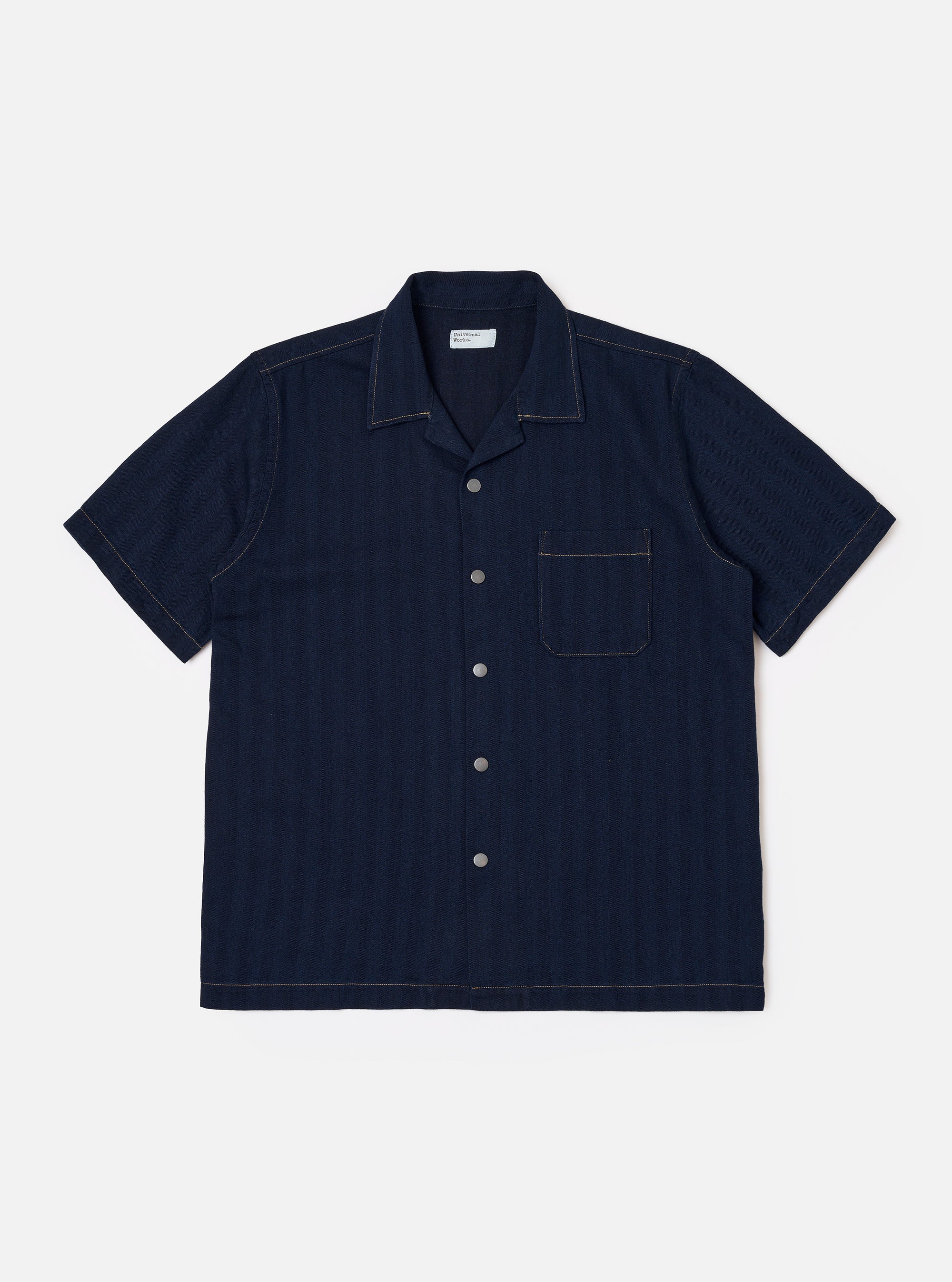 The Indigo Recycled Herringbone Road Shirt is shown as a flat lay against a white background. The short-sleeve shirt features a camp collar, a full button-front placket with brown corozo buttons, and three front patch pockets: one at the left chest and two at the waist. The fabric is made from indigo-dyed recycled cotton with a distinct herringbone weave.