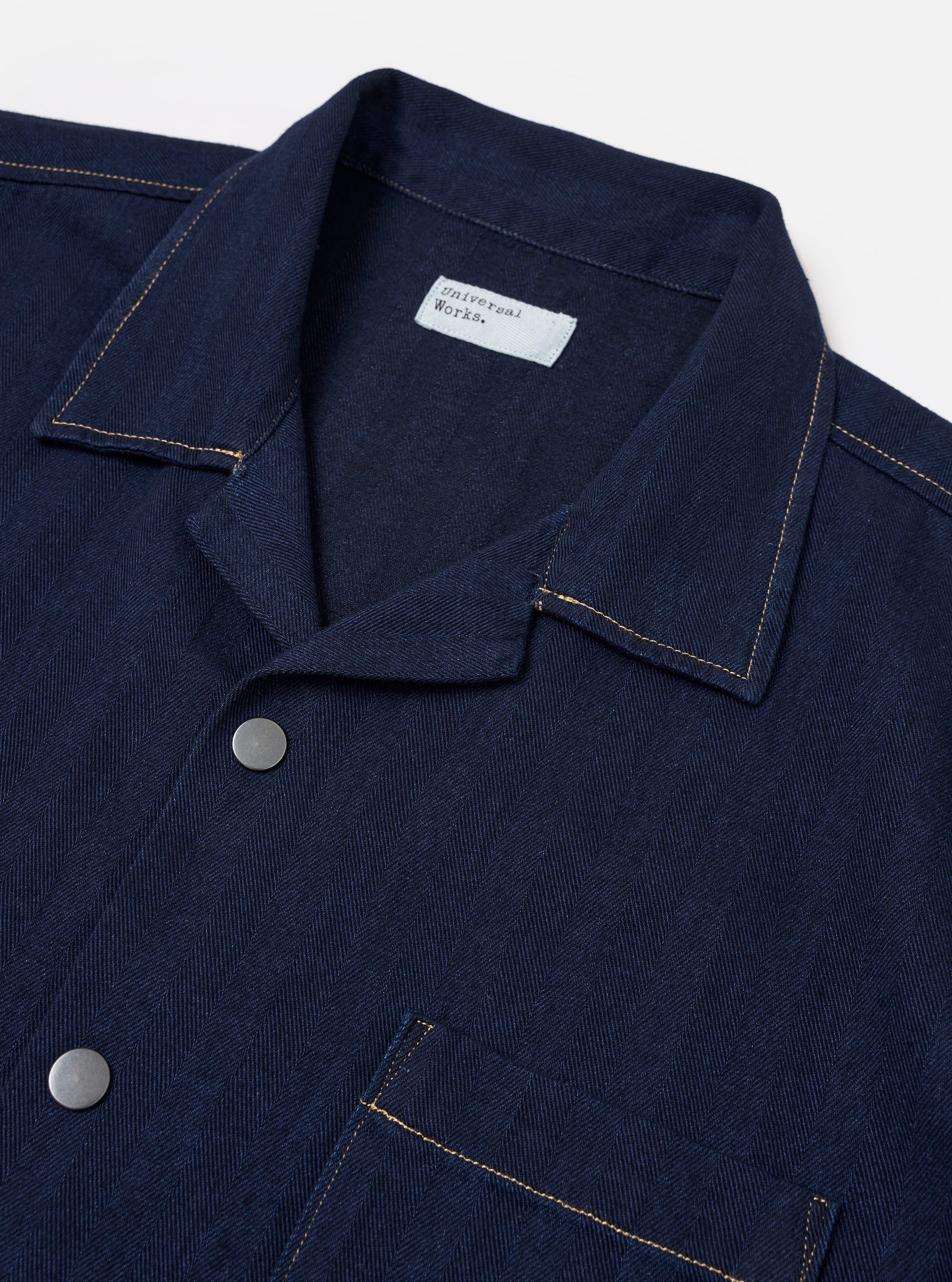 The Indigo Recycled Herringbone Road Shirt is shown from the back as a flat lay against a plain white background. The image highlights the straight hem, the back yolk, and the tonal indigo herringbone weave of the fabric.