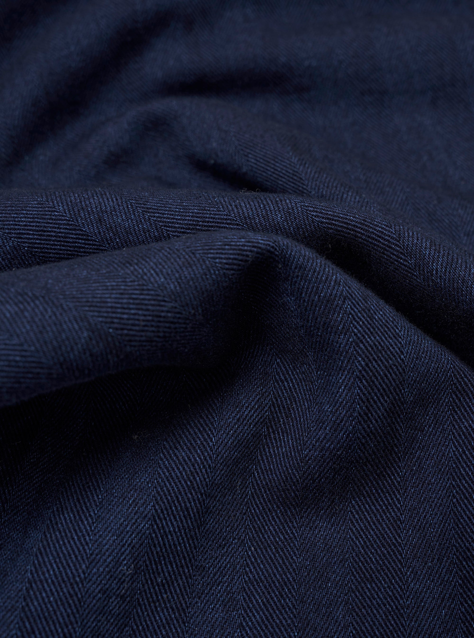 A close-up fabric swatch of the Indigo Recycled Herringbone Road Shirt displays a rich indigo blue color and a distinct V-shaped herringbone weave pattern. The image highlights the texture and tonal depth of the recycled cotton fabric.