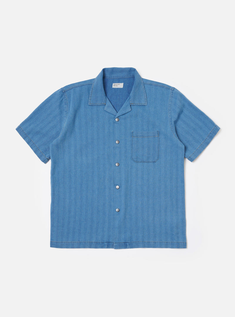 The Washed Indigo Recycled Herringbone Road Shirt is shown as a flat lay against a plain white background. The shirt features a camp collar, a six-button front closure, and two large chest pockets with button-down flaps. The fabric is a washed indigo blue with a visible herringbone weave.