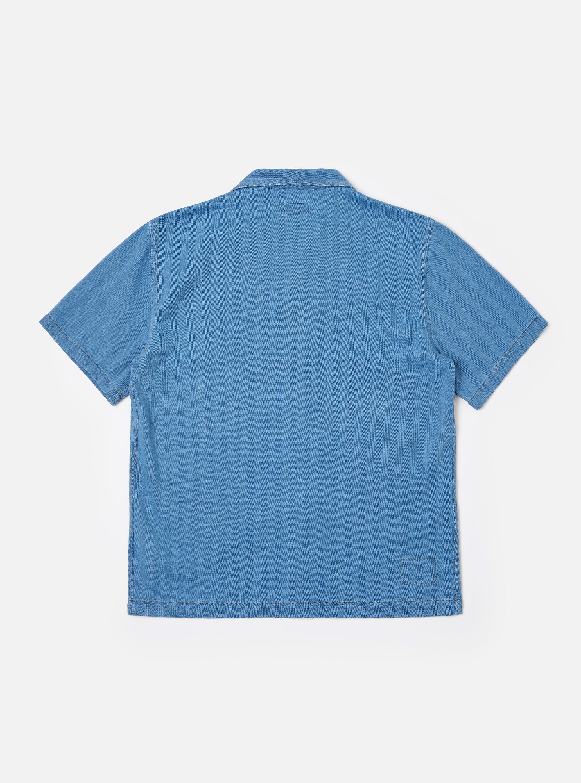 The Washed Indigo Recycled Herringbone Road Shirt is shown from the back as a flat lay against a plain white background. The image highlights the shirt's straight hem, simple collar, and the subtle chevron texture of the washed indigo herringbone fabric.