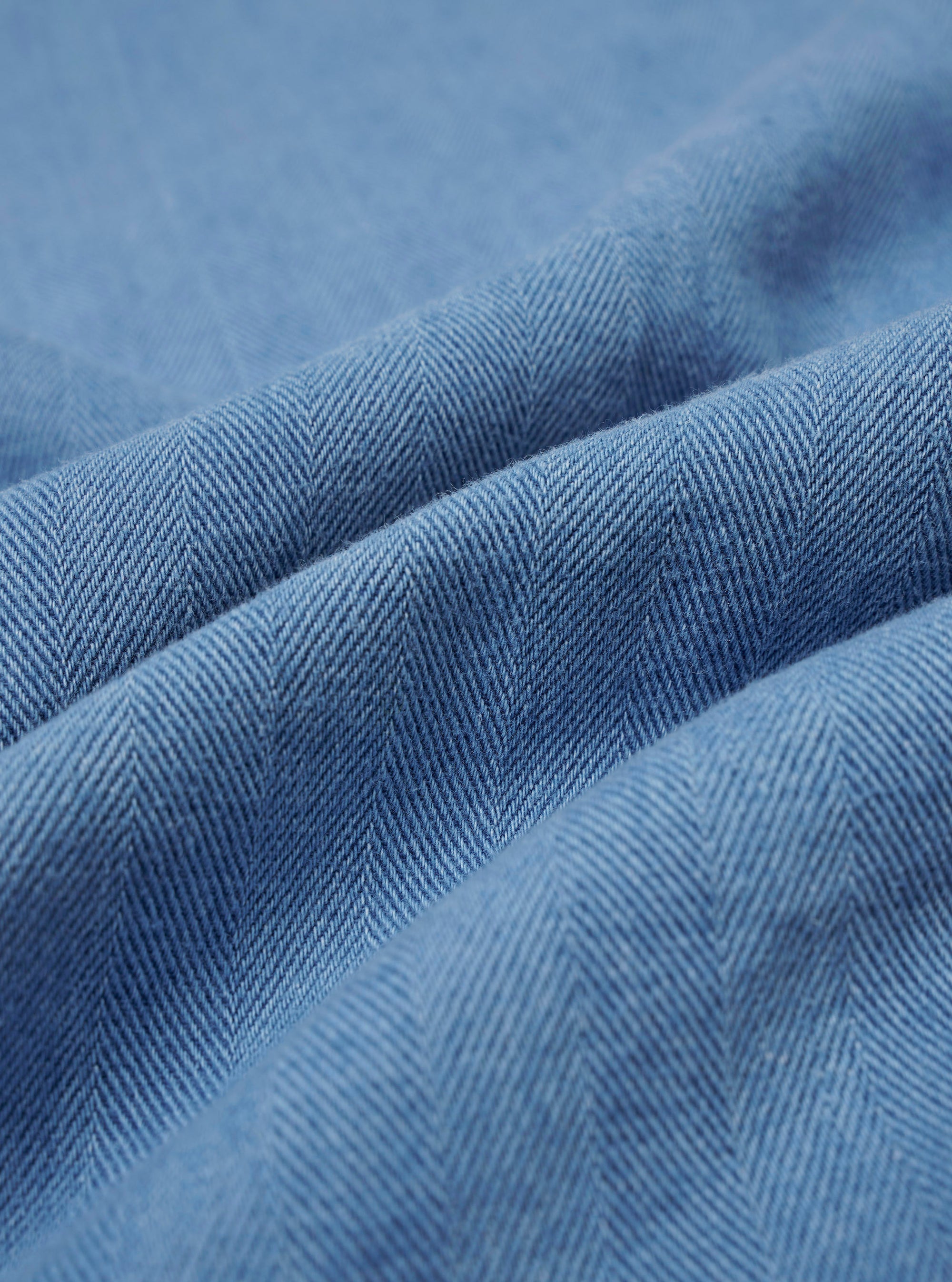 A close-up fabric swatch of the Washed Indigo Recycled Herringbone Road Shirt highlights the intricate herringbone weave pattern. The image showcases the varied blue tones of the washed indigo fabric, providing a detailed view of the textile's texture and color.