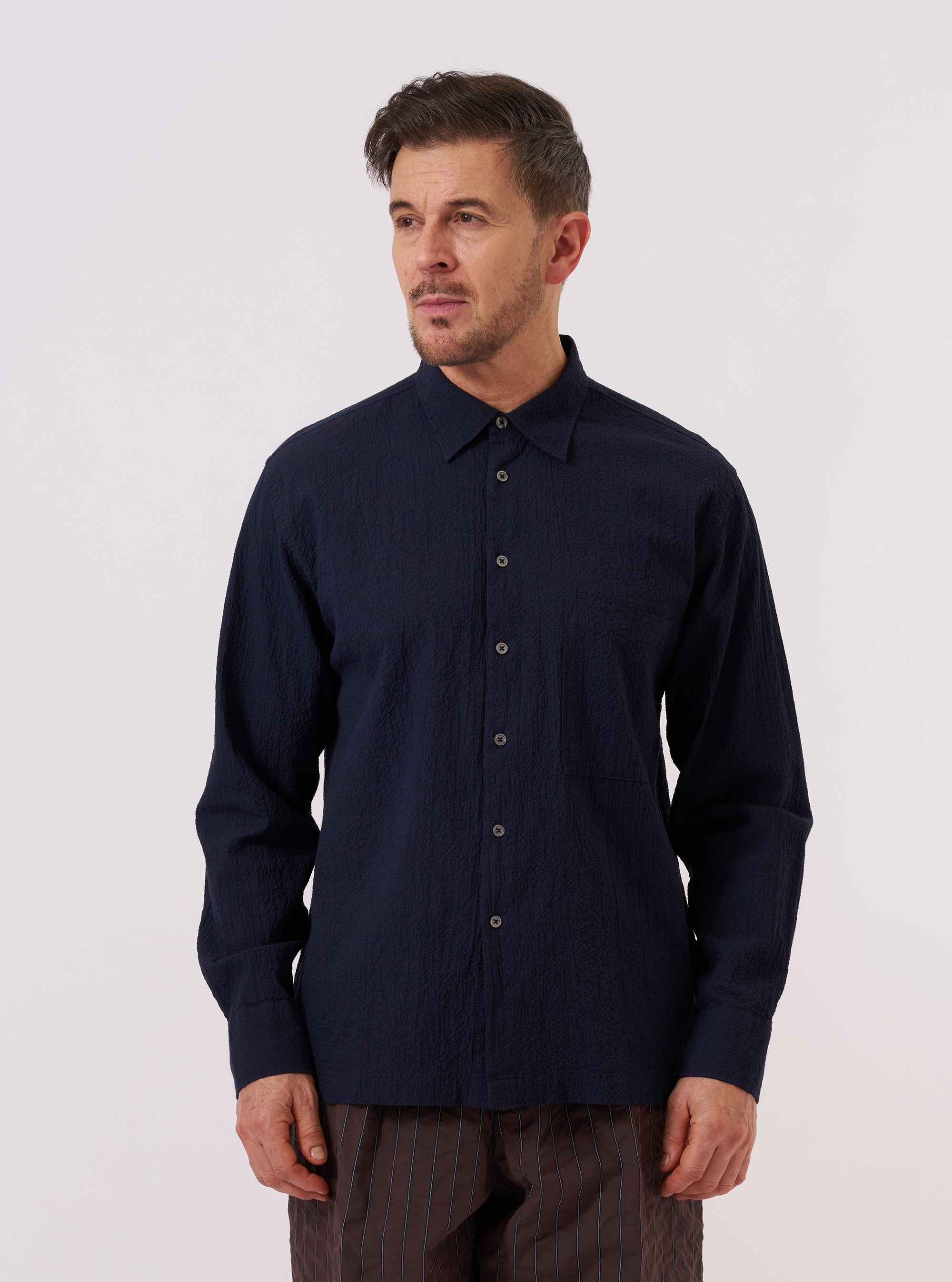 A model wears the Navy Seersucker Shirting Lucky Shirt against a plain, neutral background. The shirt features a relaxed fit with a camp collar, a central button-down closure, and short sleeves. The navy blue fabric has a distinct puckered seersucker texture throughout.