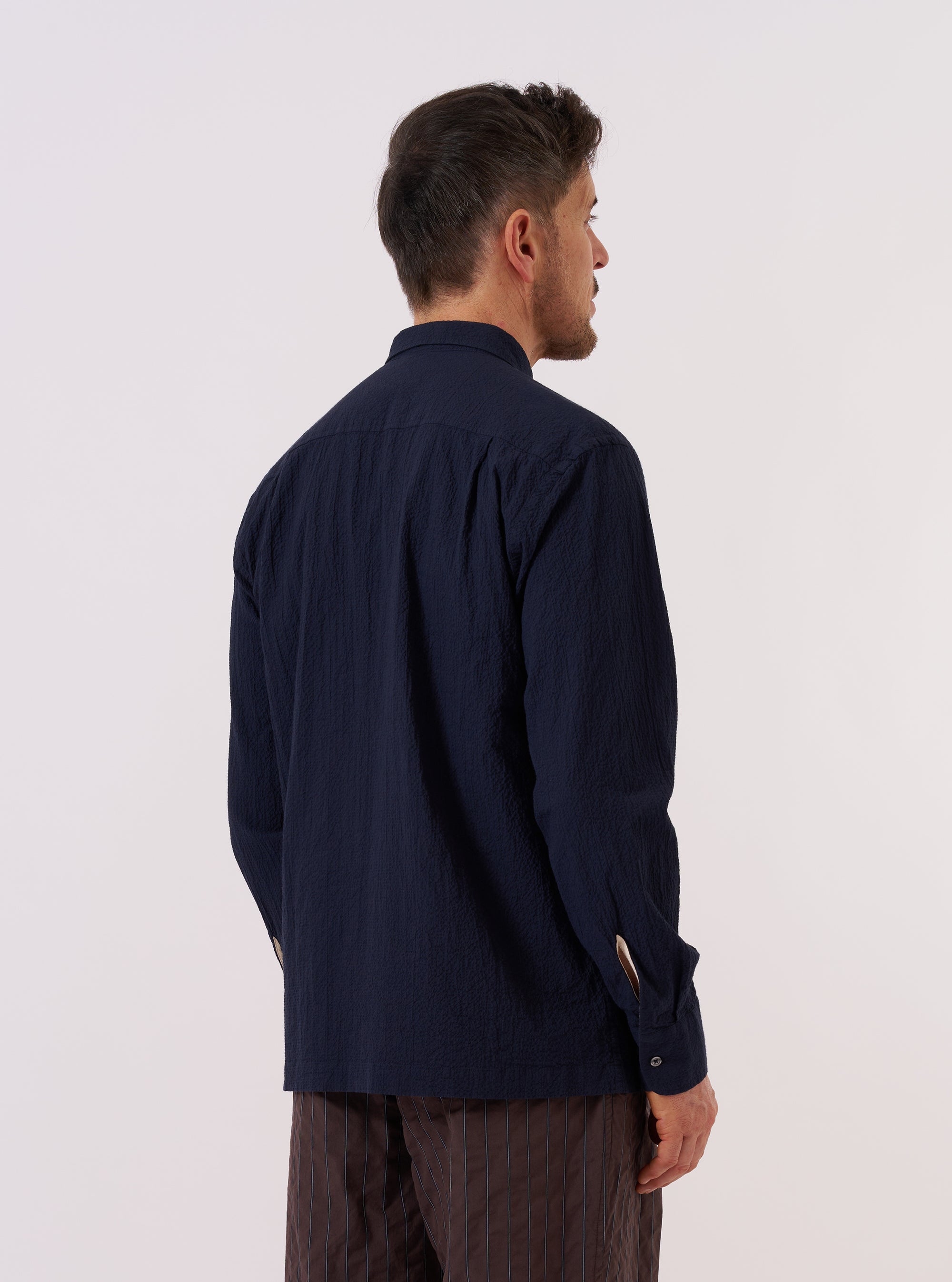 A model wears the Navy Seersucker Shirting Lucky Shirt, shown from a rear three-quarter angle against a plain, neutral background. The shirt features a relaxed fit, a straight hem, and a simple pointed collar. The navy blue cotton fabric has a distinct puckered seersucker texture throughout.