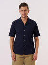 A model wears the Navy Seersucker Shirting Road Shirt against a plain, neutral background. The shirt features a relaxed camp collar, short sleeves, and a central button closure. The navy blue cotton fabric displays a distinct, puckered seersucker texture throughout. The model is shown from the waist up, wearing the shirt untucked.