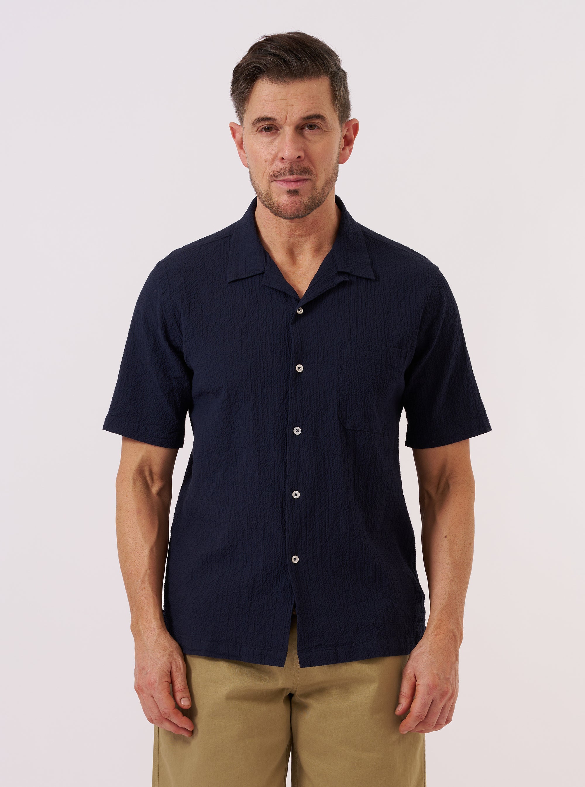A model wears the Navy Seersucker Shirting Road Shirt against a plain, neutral background. The shirt features a relaxed camp collar, short sleeves, and a central button closure. The navy blue cotton fabric displays a distinct, puckered seersucker texture throughout. The model is shown from the waist up, wearing the shirt untucked.
