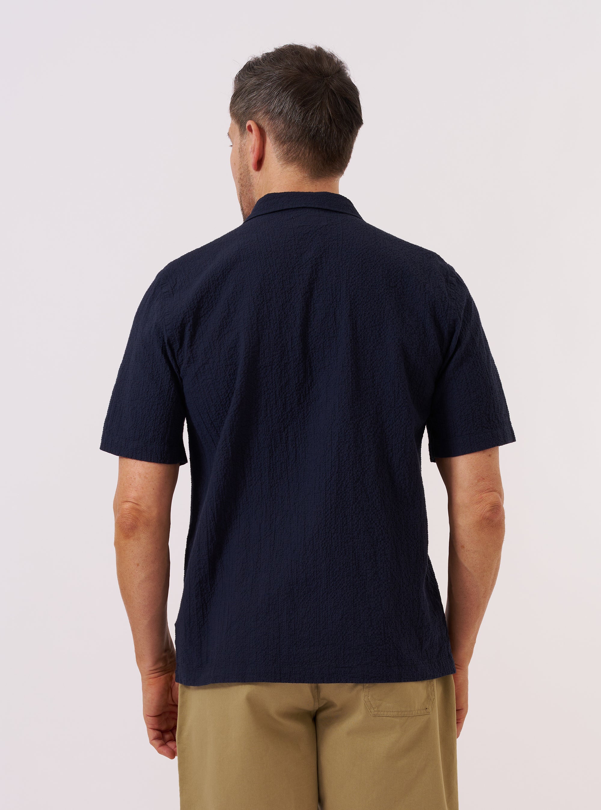 A model wears the Navy Seersucker Shirting Road Shirt, shown from the back against a plain white background. The shirt features a relaxed fit, short sleeves, and a straight hem. The dark navy fabric has a distinct puckered seersucker texture. The model is wearing the shirt tucked into light-colored trousers.