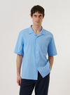 The Sky Blue Seersucker Shirting Road Shirt is shown as a flat lay against a plain white background. The shirt features a camp collar, a central button-down front with white buttons, and a single patch pocket on the left chest. The short-sleeved garment is made from a light blue and white striped seersucker fabric with a visible puckered texture.
