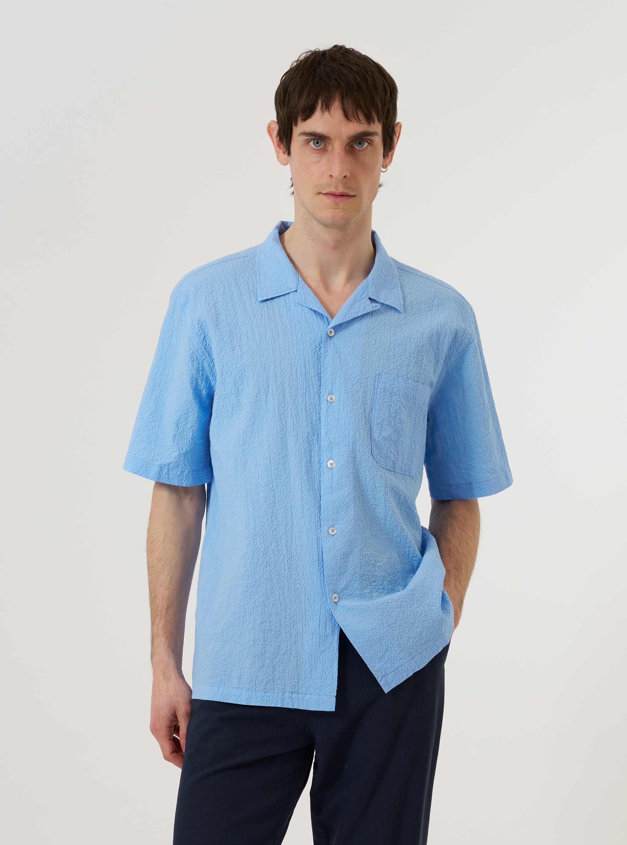 The Sky Blue Seersucker Shirting Road Shirt is shown as a flat lay against a plain white background. The shirt features a camp collar, a central button-down front with white buttons, and a single patch pocket on the left chest. The short-sleeved garment is made from a light blue and white striped seersucker fabric with a visible puckered texture.