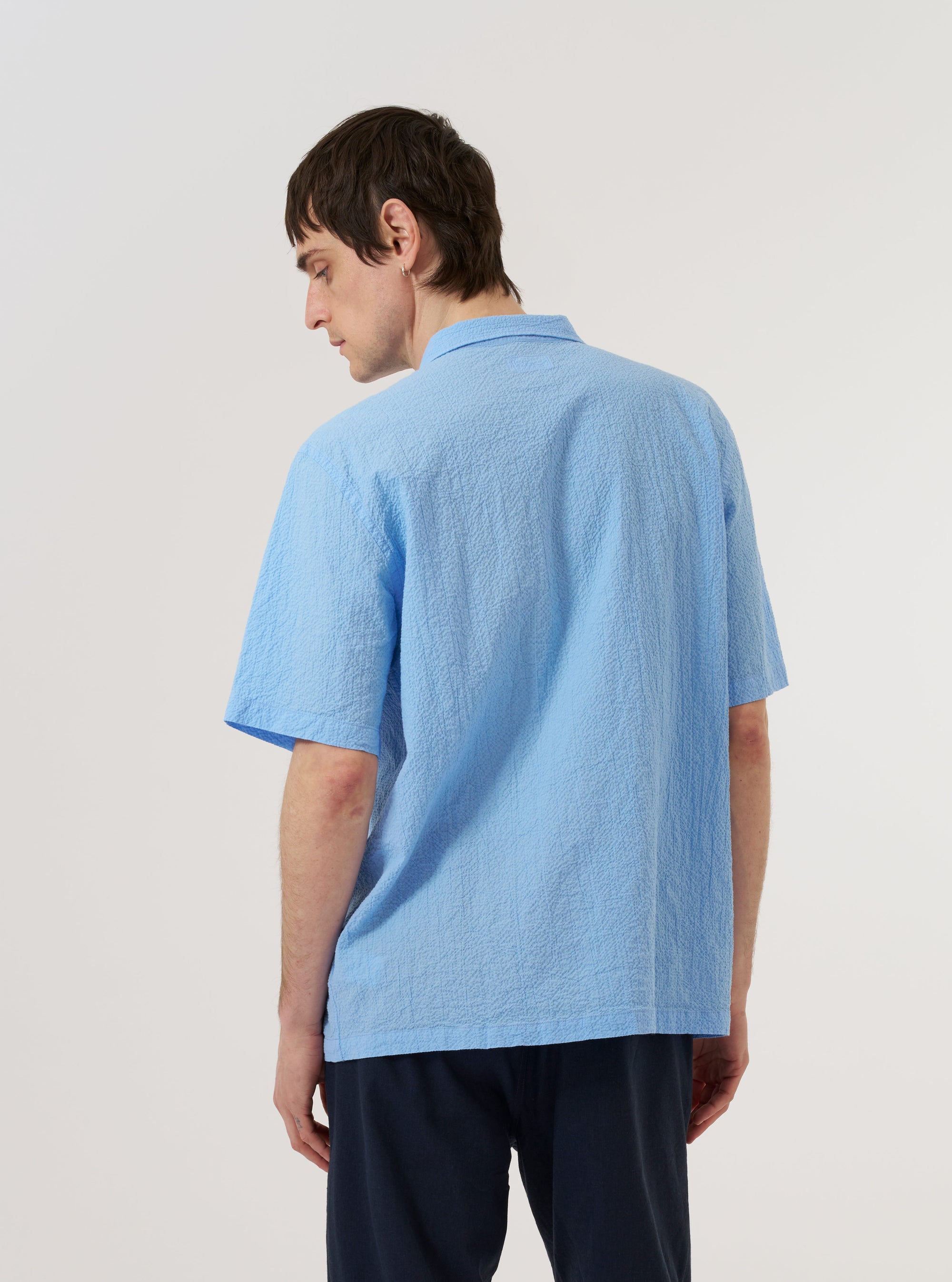 The Sky Blue Seersucker Shirting Road Shirt is shown from the back as a flat lay against a plain white background. The image highlights the shirt's straight hem, simple back yoke, and the distinct puckered texture of the sky blue seersucker fabric.