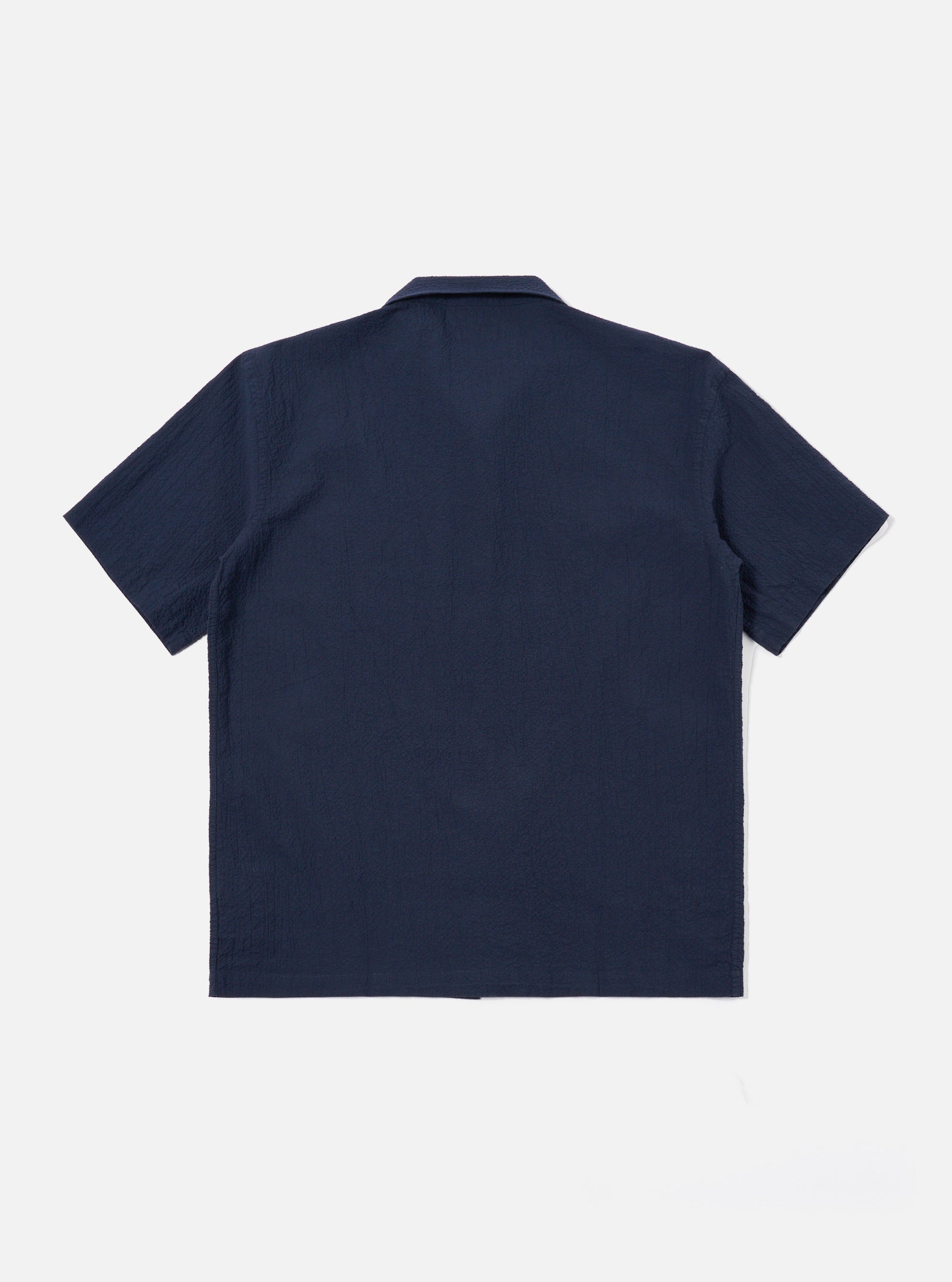 The Navy Seersucker Shirting Road Shirt is shown from a rear angle as a flat lay against a plain white background. The image highlights the shirt's short sleeves, straight hem, and the subtle puckered texture of the navy blue seersucker fabric. A small locker loop is visible at the center of the back yoke.