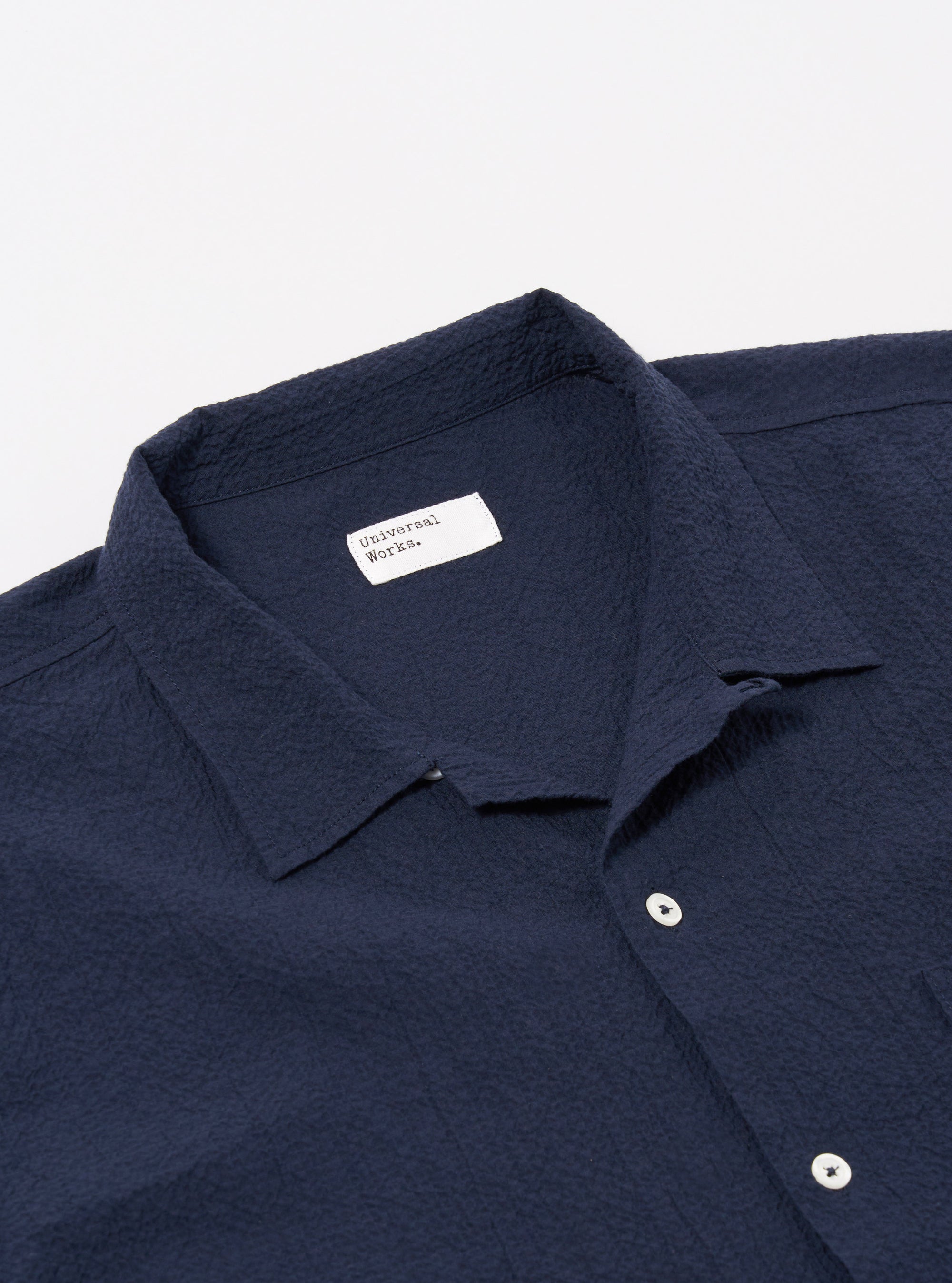 The Navy Seersucker Shirting Road Shirt is shown from the back as a flat lay against a plain white background. The image highlights the shirt's straight hem, short sleeves, and the distinct puckered texture of the navy blue seersucker fabric. A horizontal yoke across the shoulders and a simple, clean construction are visible.