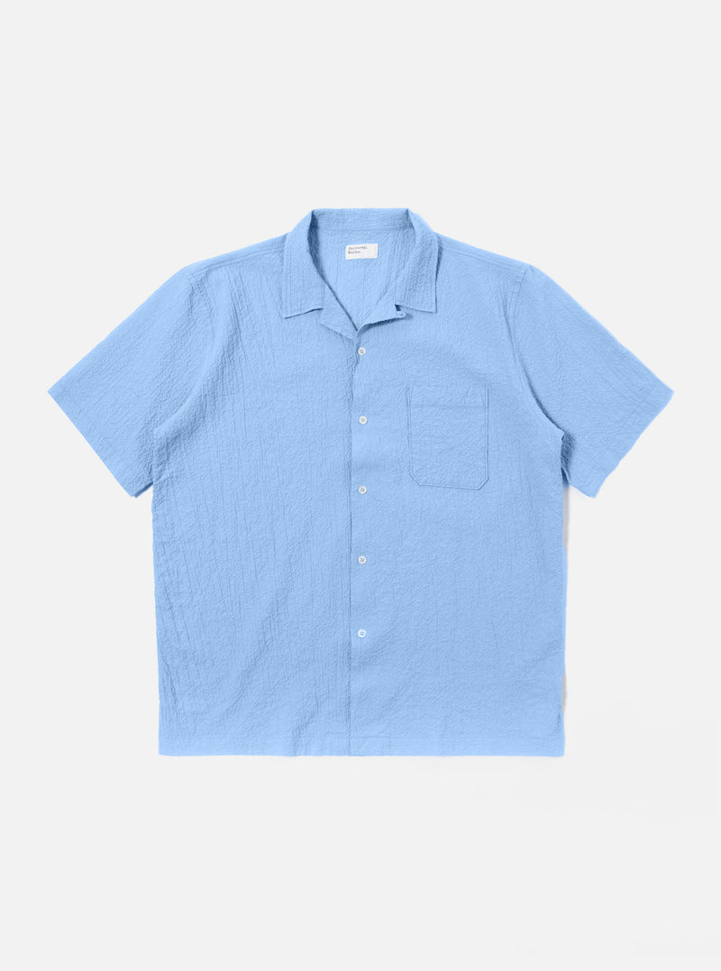 The Sky Blue Seersucker Shirting Road Shirt is shown as a flat lay against a plain white background. The short-sleeve shirt features a camp collar with a loop closure, a single chest patch pocket, and a straight hem. The fabric has a visible puckered seersucker texture in a solid sky blue color.