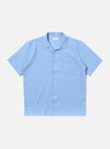 The Sky Blue Seersucker Shirting Road Shirt is shown as a flat lay against a plain white background. The short-sleeve shirt features a camp collar with a loop closure, a single chest patch pocket, and a straight hem. The fabric has a visible puckered seersucker texture in a solid sky blue color.
