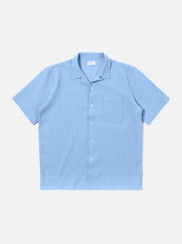 The Sky Blue Seersucker Shirting Road Shirt is shown as a flat lay against a plain white background. The short-sleeve shirt features a camp collar with a loop closure, a single chest patch pocket, and a straight hem. The fabric has a visible puckered seersucker texture in a solid sky blue color.