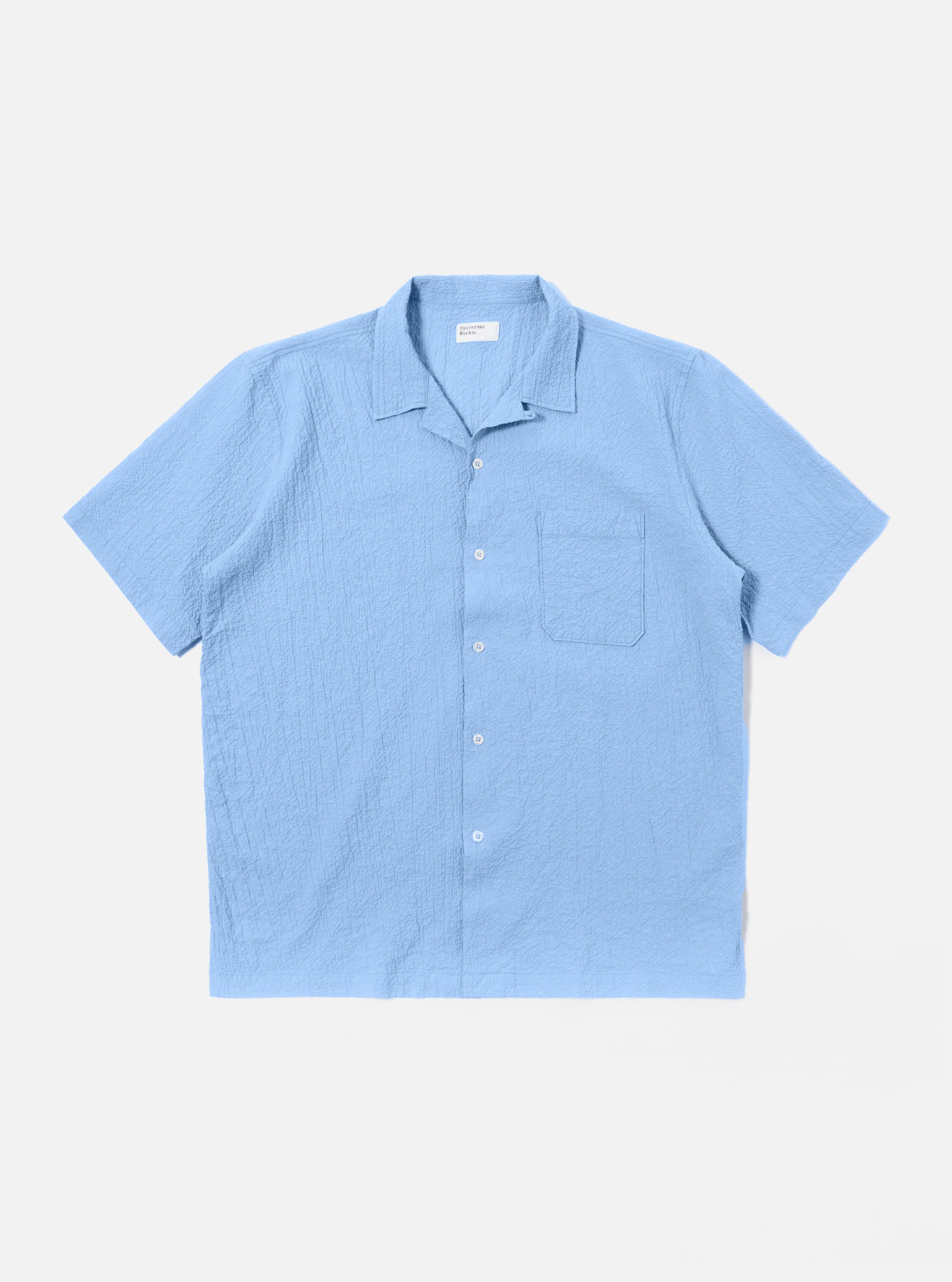 The Sky Blue Seersucker Shirting Road Shirt is shown as a flat lay against a plain white background. The short-sleeve shirt features a camp collar with a loop closure, a single chest patch pocket, and a straight hem. The fabric has a visible puckered seersucker texture in a solid sky blue color.