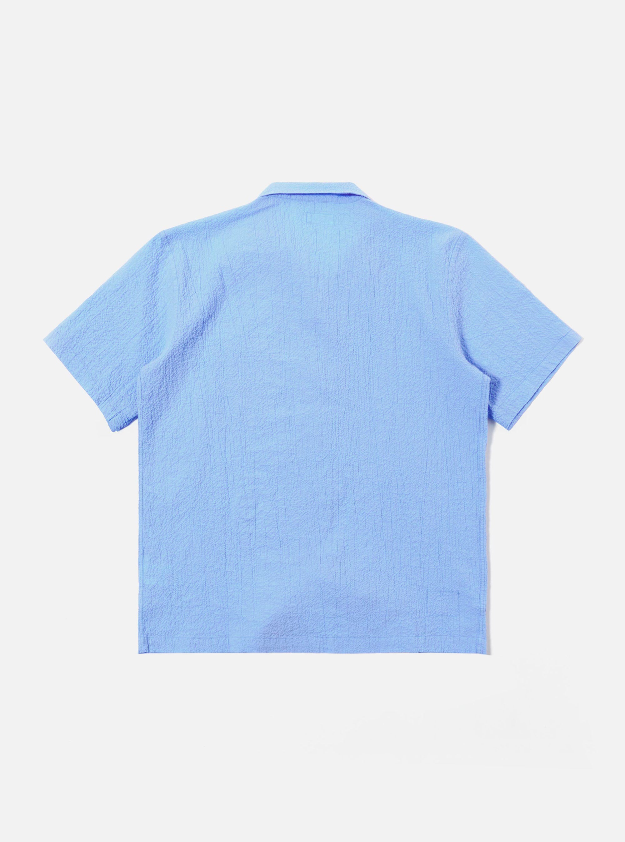 The Sky Blue Seersucker Shirting Road Shirt is shown from the back as a flat lay against a plain white background. The image highlights the shirt's short sleeves, straight hem, and the puckered texture of the sky blue seersucker fabric.