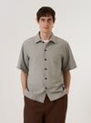 A model wears the Grey Izumi Cotton S/S Fatigue Shirt against a plain, neutral background. The shirt features two large patch pockets on the chest, a central button-down closure, and a structured collar. The grey cotton fabric has a visible fine texture. The model is wearing the short-sleeved shirt over a light-colored top.
