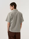 A model wears the Grey Izumi Cotton S/S Fatigue Shirt, shown from a rear three-quarter angle against a plain white background. The short-sleeve shirt features a straight hem, a structured collar, and a clean back panel. The grey cotton fabric displays a subtle, textured weave.