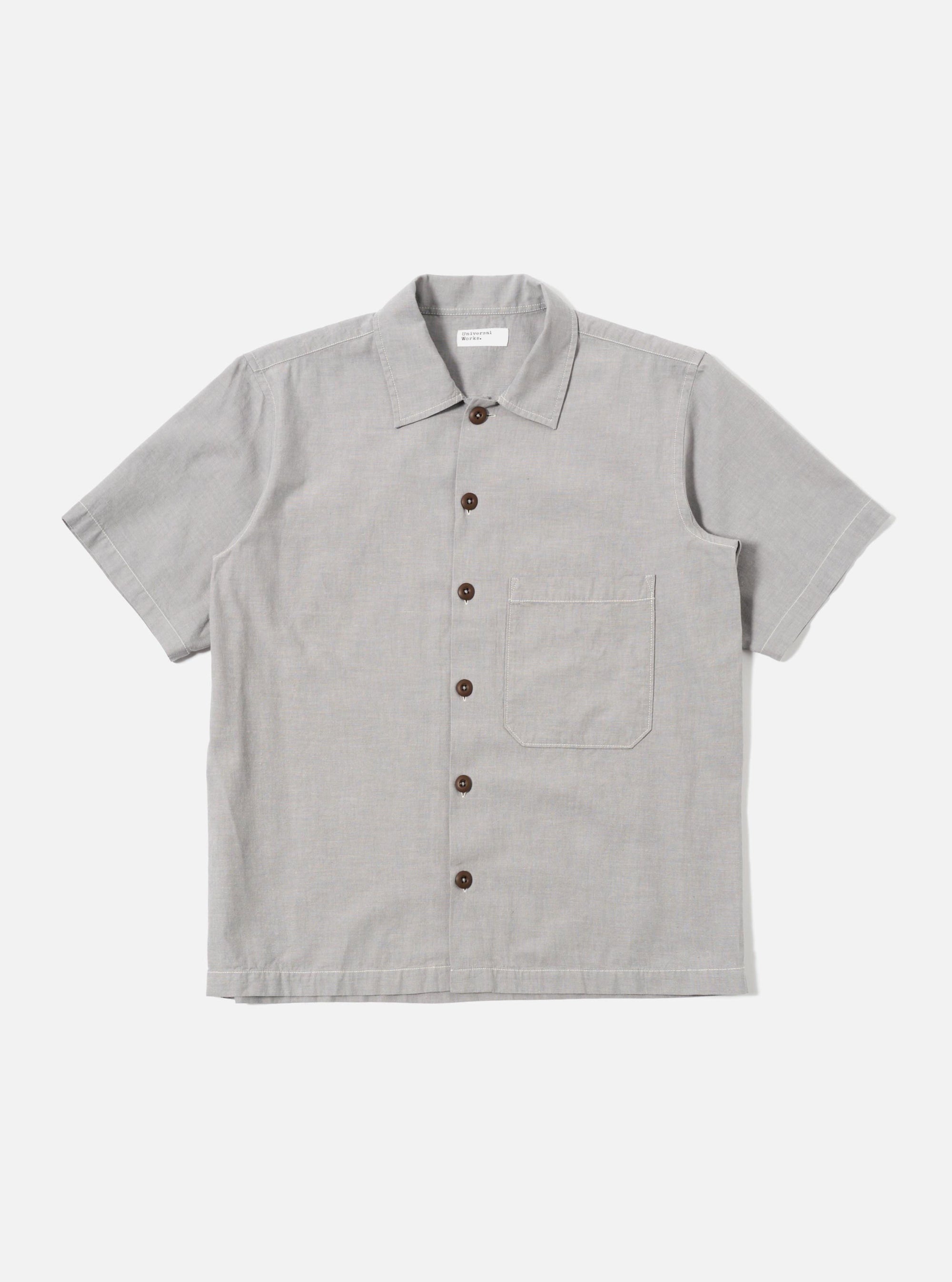 The Grey Izumi Cotton S/S Fatigue Shirt is shown as a flat lay against a white background. The shirt features a classic collar, a central button-down closure, and short sleeves. Two large patch pockets with button flaps are prominent on the chest. The fabric is a solid grey cotton with a smooth texture.