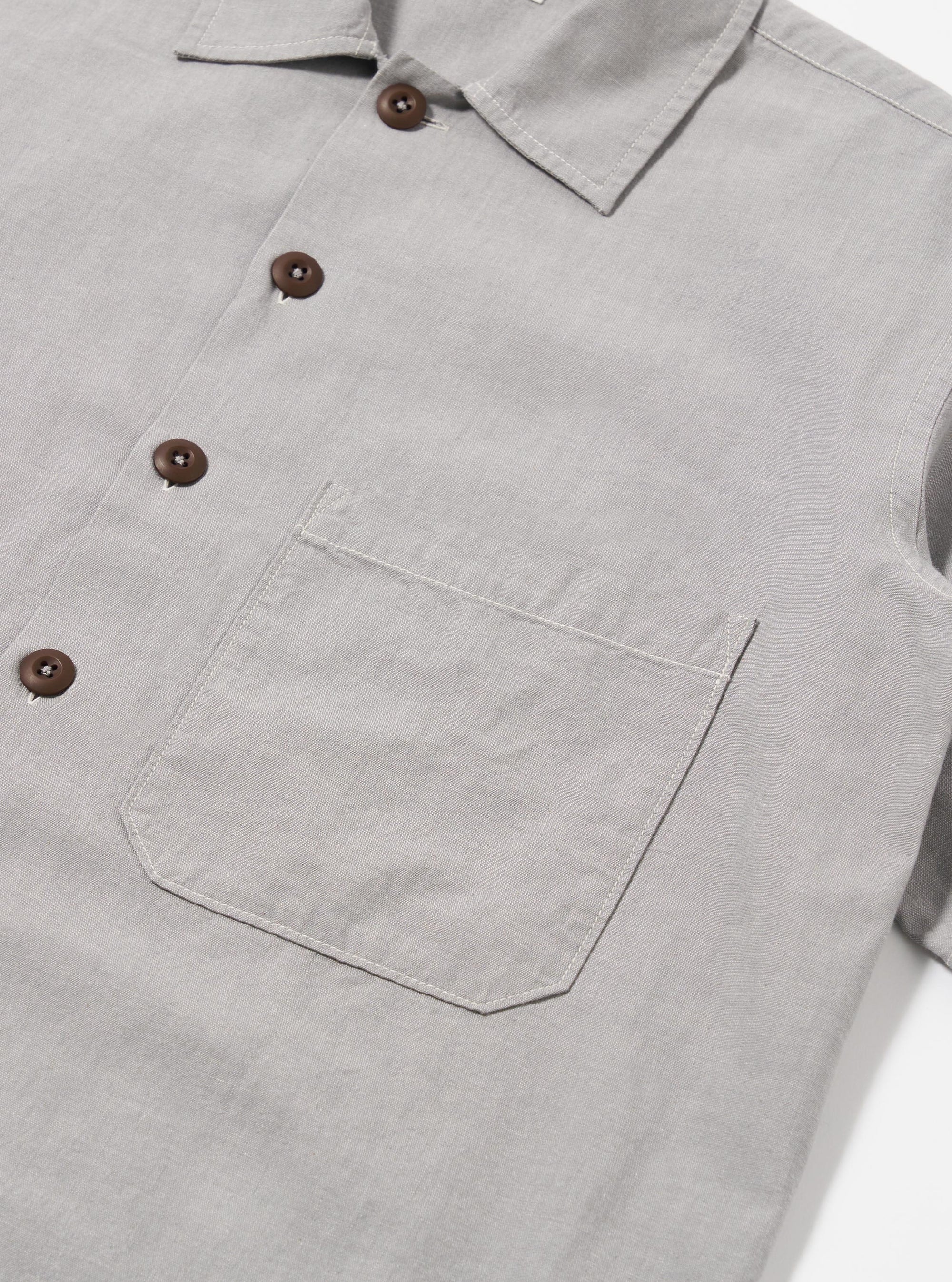 The Grey Izumi Cotton S/S Fatigue Shirt is shown in a detail view as a flat lay against a plain white background. The image highlights the textured, slubby character of the grey cotton-linen fabric and the contrasting white stitch detailing along the collar and seams.