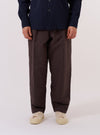 A model wears the Brown Chester Stripe Oxford Pant, standing against a plain white background. The trousers feature a loose, wide-leg fit with a single pleat at the front and a brown vertical stripe pattern on a lighter base. The model is shown from the waist down, paired with dark shoes, highlighting the garment's tapered silhouette and mid-height rise.