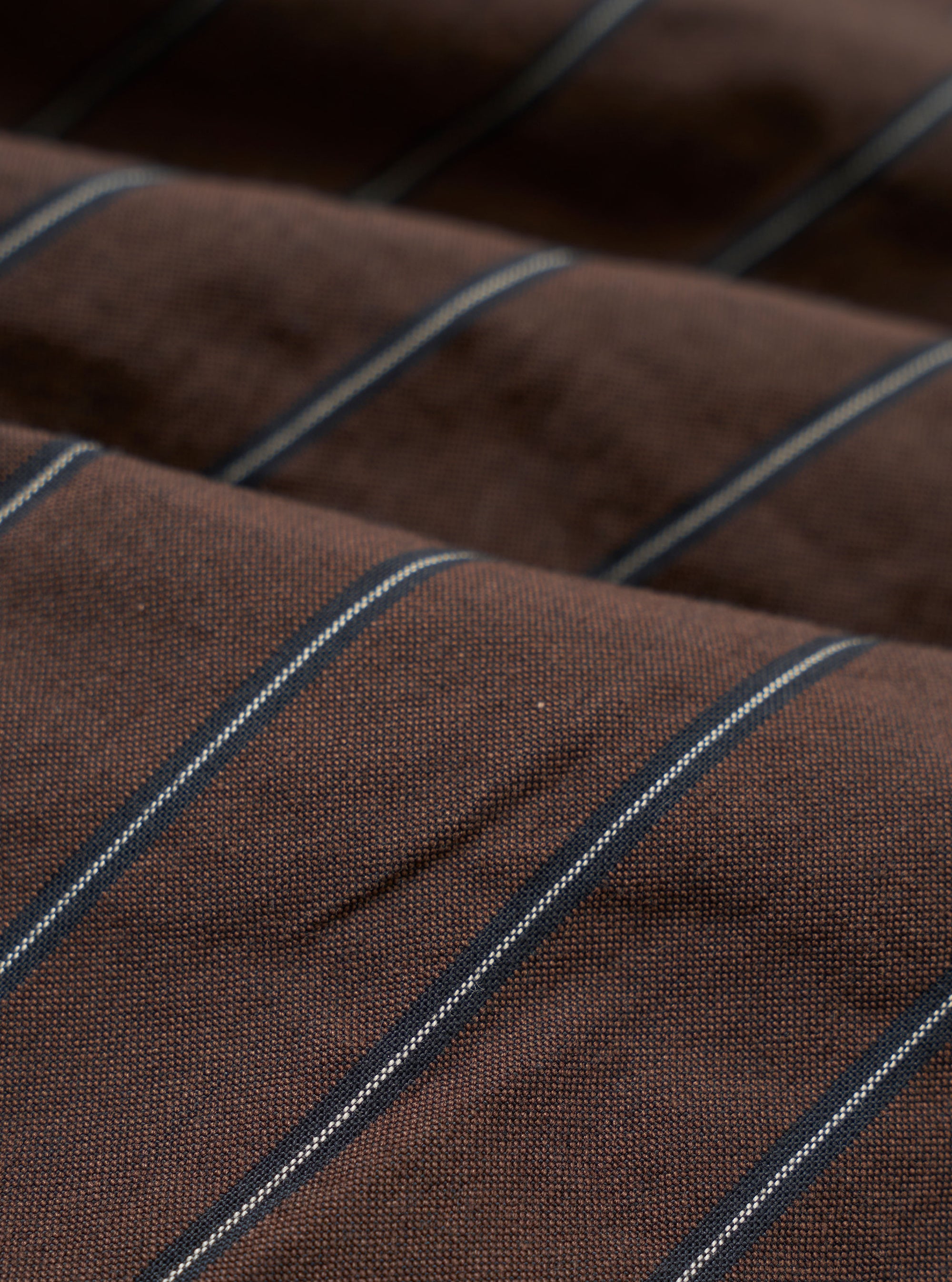 A close-up fabric swatch of the Brown Chester Stripe Oxford Pant highlights the texture and pattern of the material. The image displays a brown base with thin, light-colored vertical stripes across the cotton fabric.