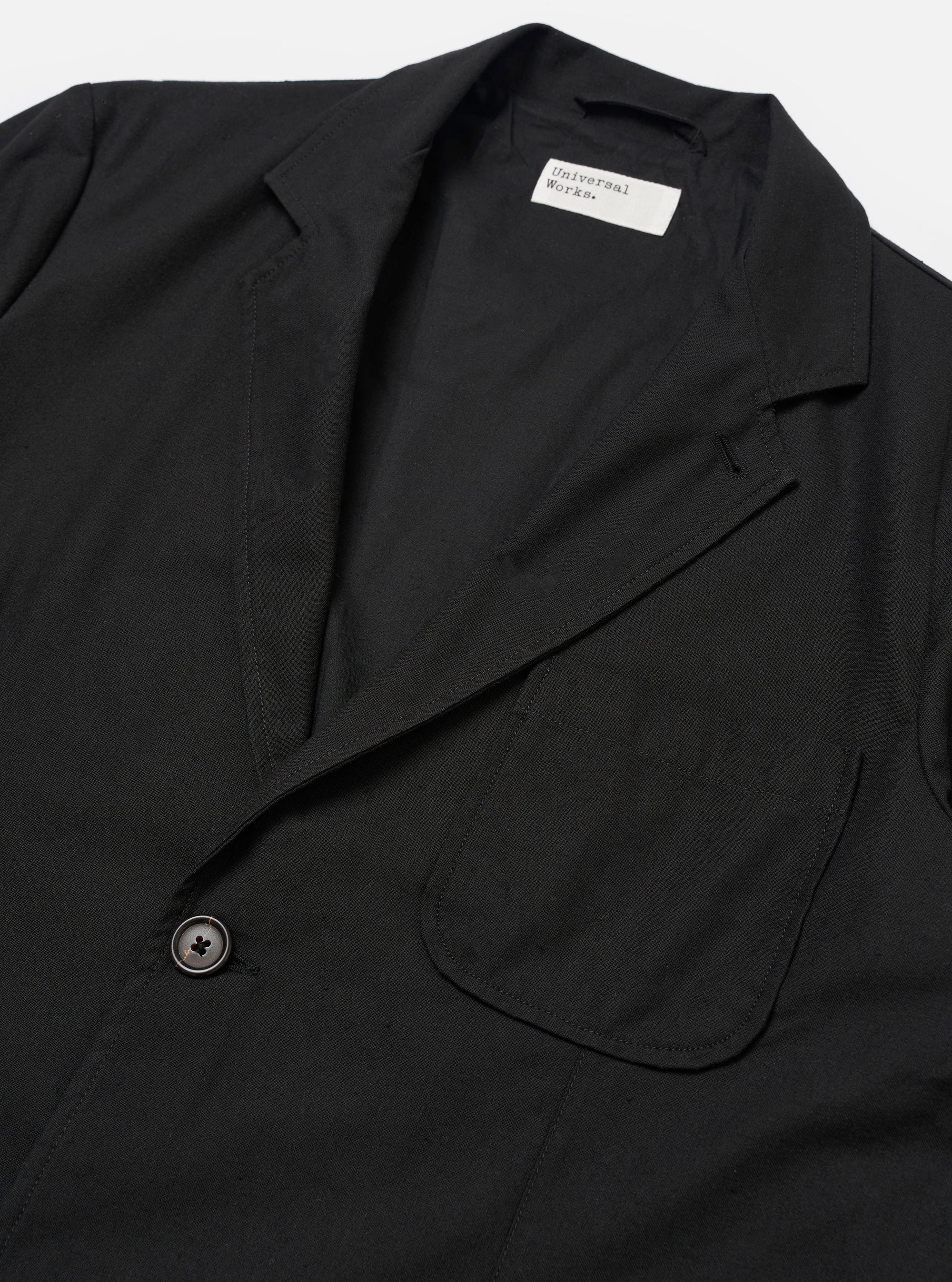 The Black Valencia Cotton Two Button Jacket is worn by a model against a plain white background. The image shows a rear three-quarter view, highlighting the jacket's loose fit, structured shoulders, and central back seam. The black fabric has a smooth finish, and the model is wearing the jacket over a light-colored long-sleeve top. No mannequin is present in the image.