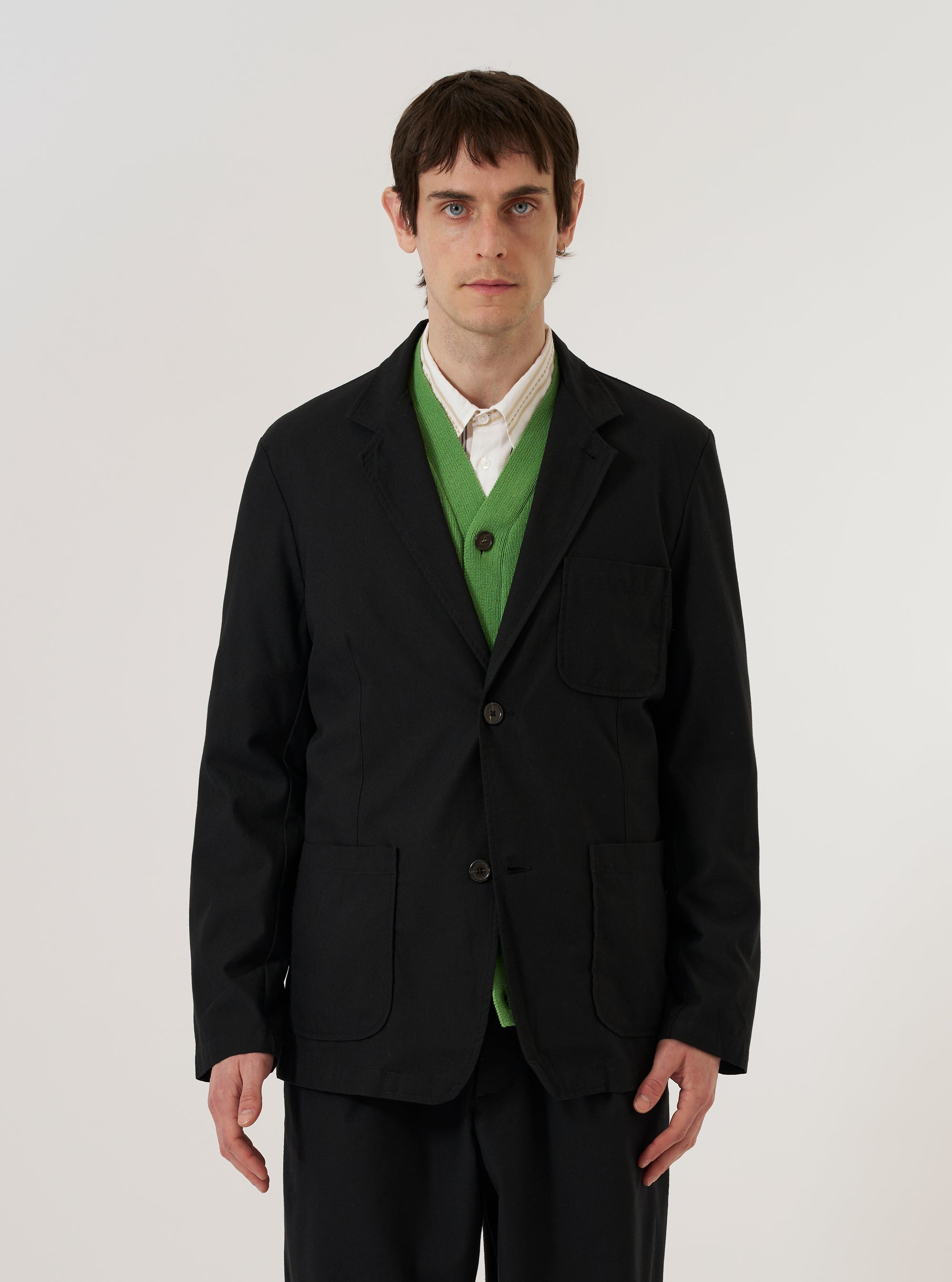A model wears the Black Valencia Cotton Two Button Jacket over a light-colored shirt, standing against a plain, neutral background. The jacket features a classic two-button front closure, notched lapels, and three front patch pockets. The black fabric has a subtle, textured appearance.