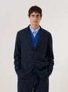 A model wears the Navy Valencia Cotton Two Button Jacket over a light-colored shirt, standing against a plain white background. The jacket features a classic notch lapel, a two-button front closure, and two large patch pockets at the waist. The navy fabric has a smooth, structured appearance suitable for suiting.