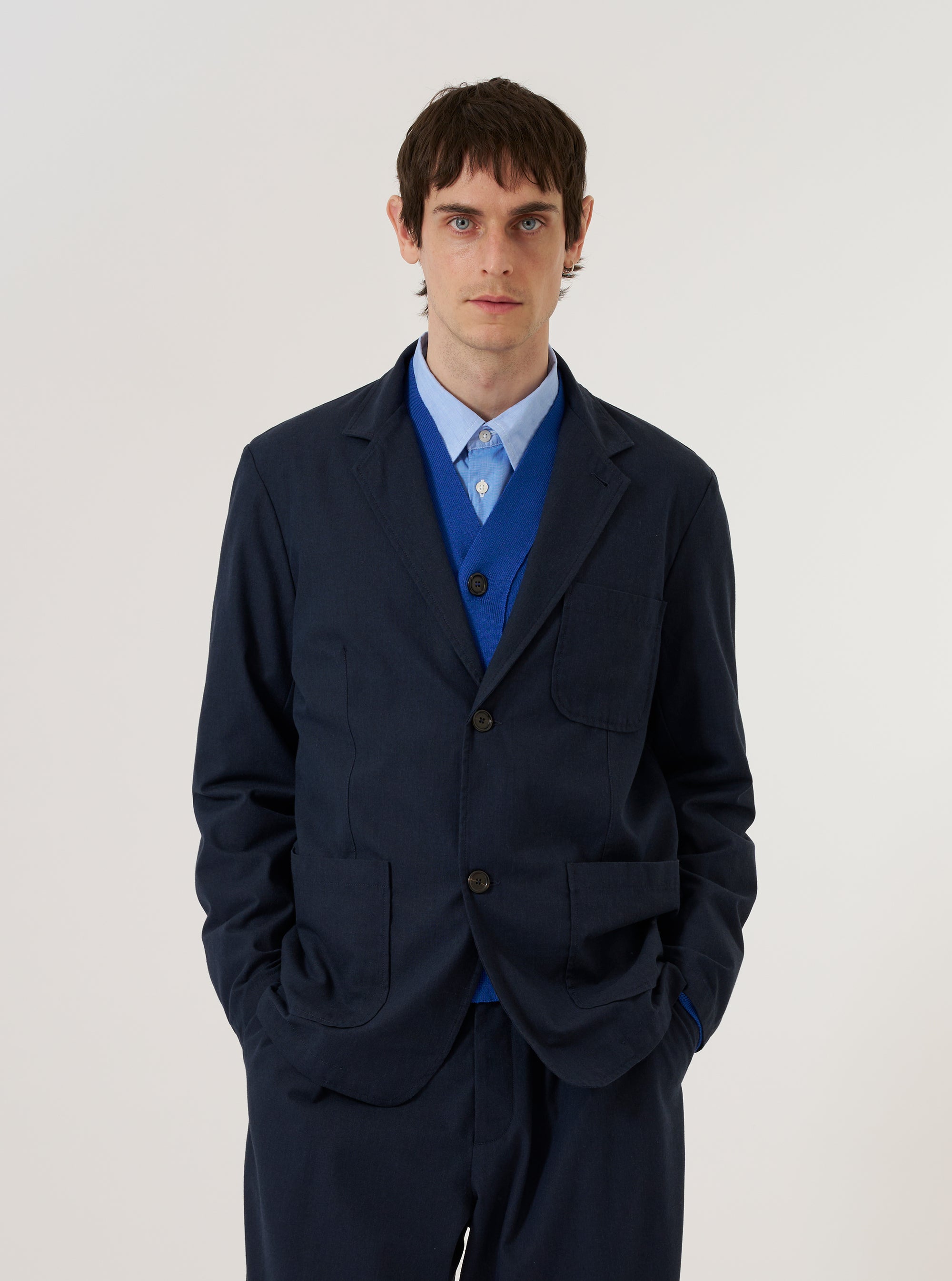 A model wears the Navy Valencia Cotton Two Button Jacket over a light-colored shirt, standing against a plain white background. The jacket features a classic notch lapel, a two-button front closure, and two large patch pockets at the waist. The navy fabric has a smooth, structured appearance suitable for suiting.