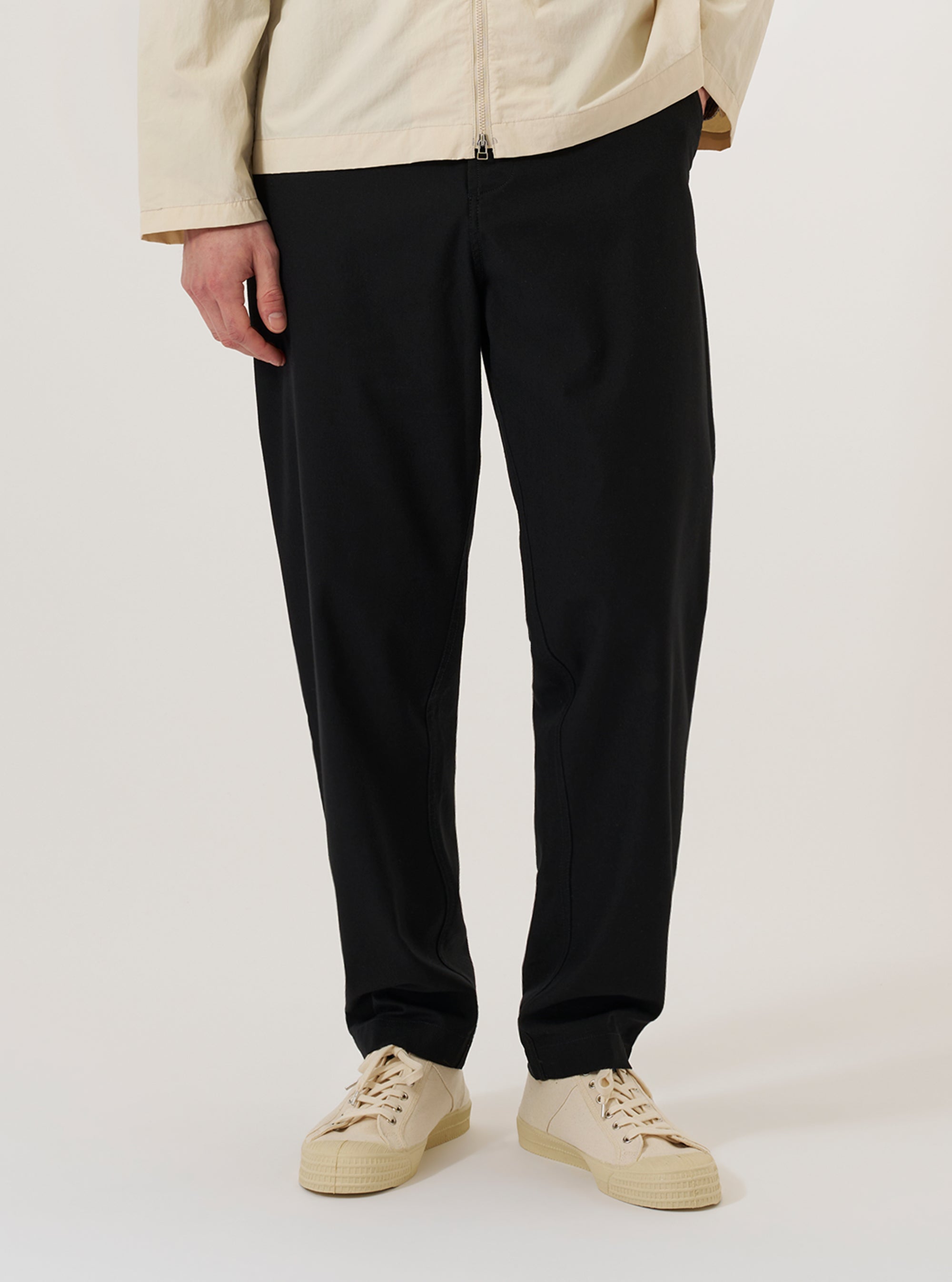 A model wears the Black Valencia Cotton Military Chino while standing against a plain white background. The trousers feature a straight-leg fit, front slash pockets, and a structured waistband with belt loops. The black fabric has a smooth, durable finish characteristic of the Valencia cotton blend. The model is shown from the waist down, highlighting the garment's classic military-inspired silhouette and clean construction.